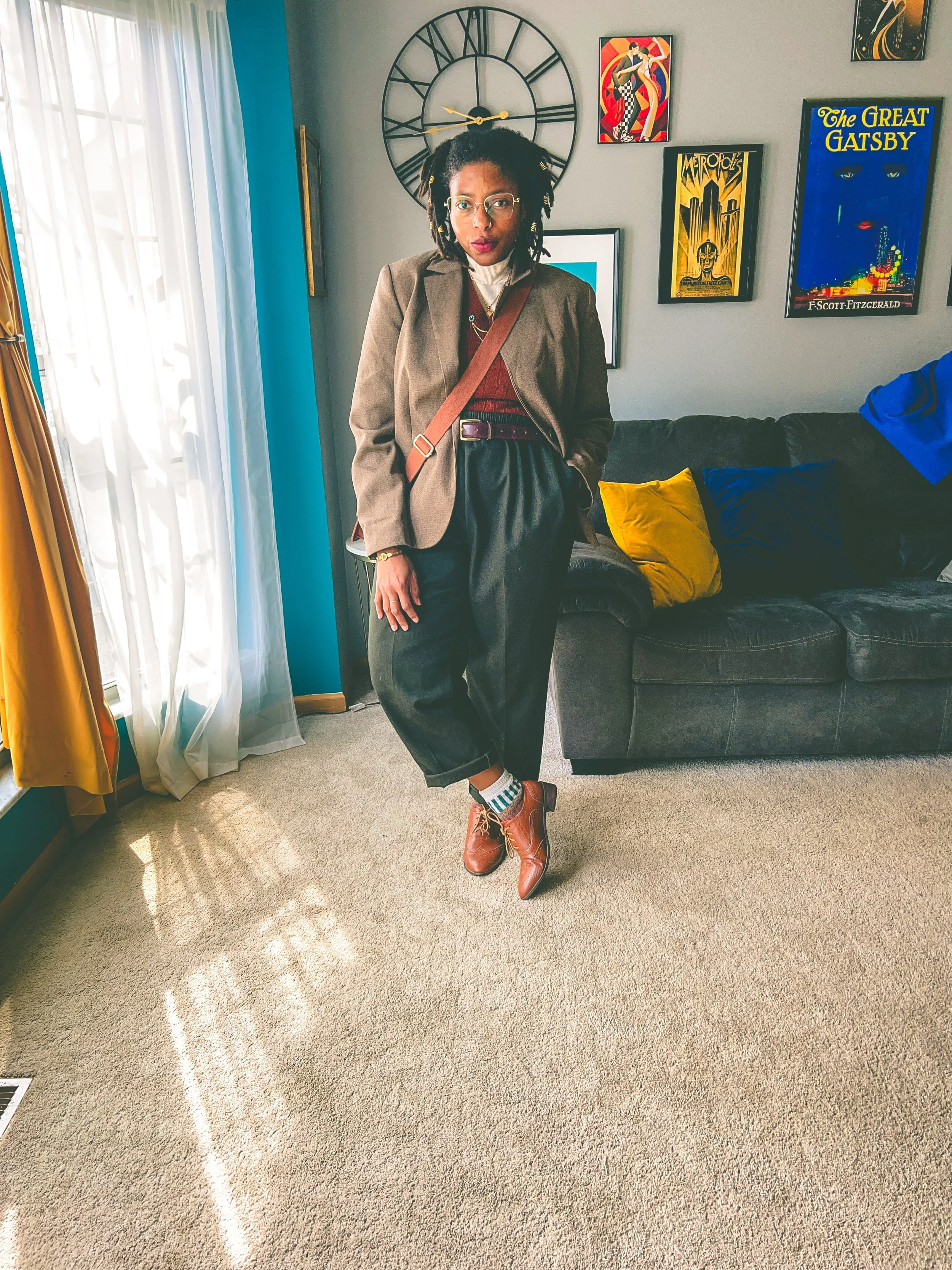A woman with glasses standing in a living room, wearing a brown blazer, red sweater, high-waisted black pants, striped socks, and brown shoes, with wall art including a clock and posters behind her.