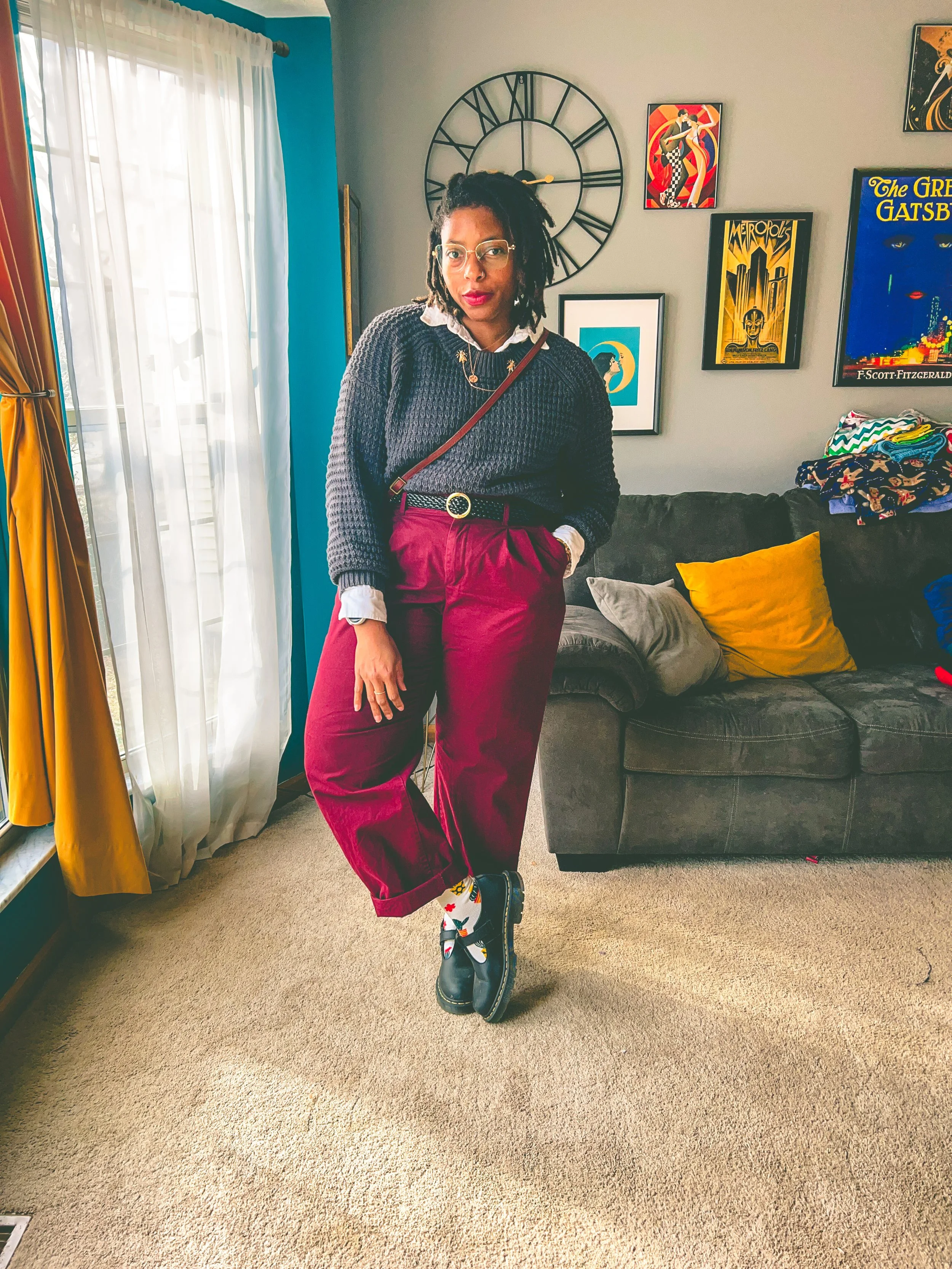 A woman standing in a living room with her hand in her pocket, wearing glasses, a black sweater, burgundy pants, and colorful socks with black boots. There is a large wall clock and framed artwork behind her, and a dark gray couch with yellow and gra