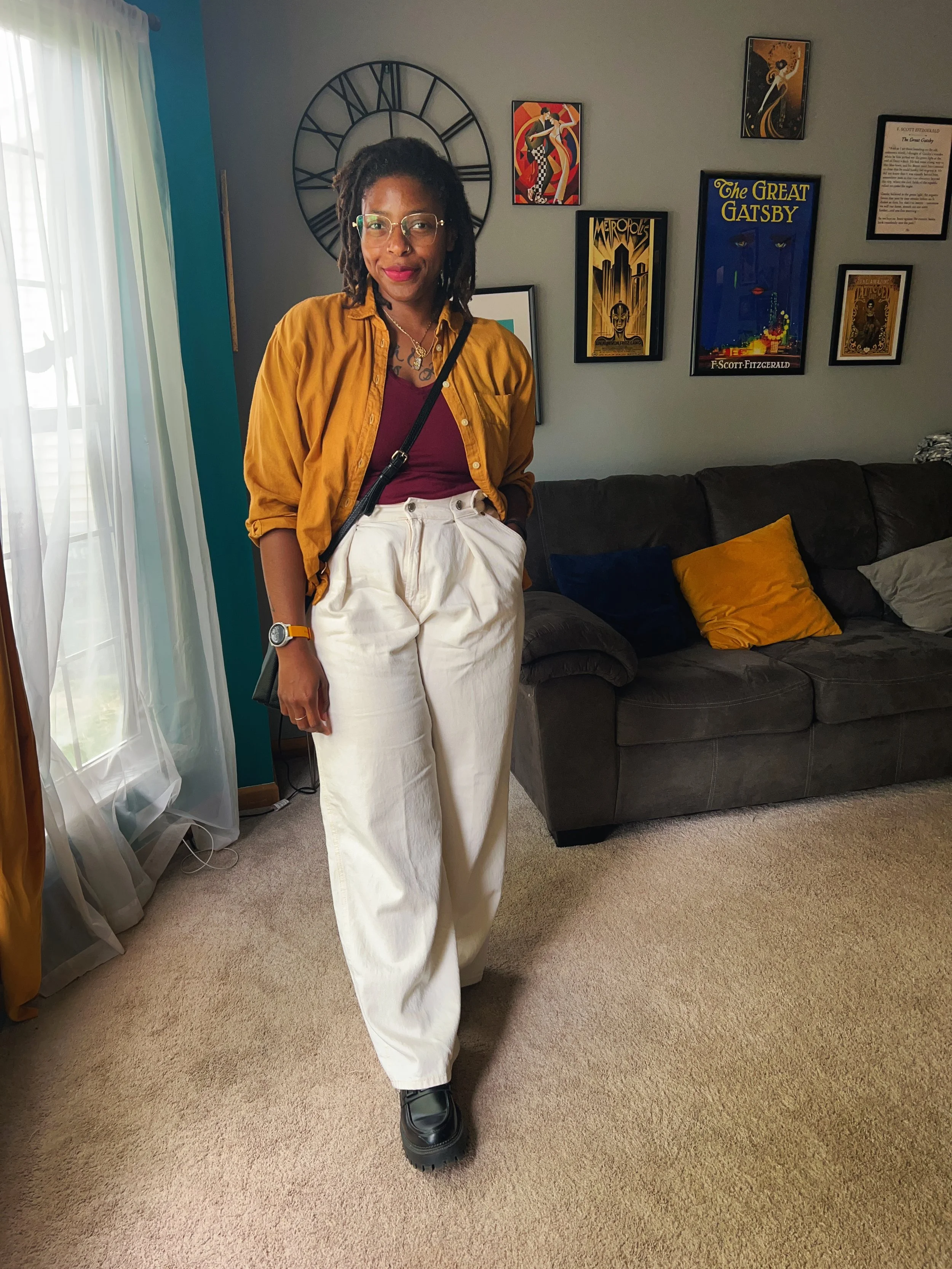 A woman with glasses and dreadlocks standing inside a living room, wearing a mustard jacket, maroon top, white pants, and black boots, with framed artwork and a large clock on the wall behind her.