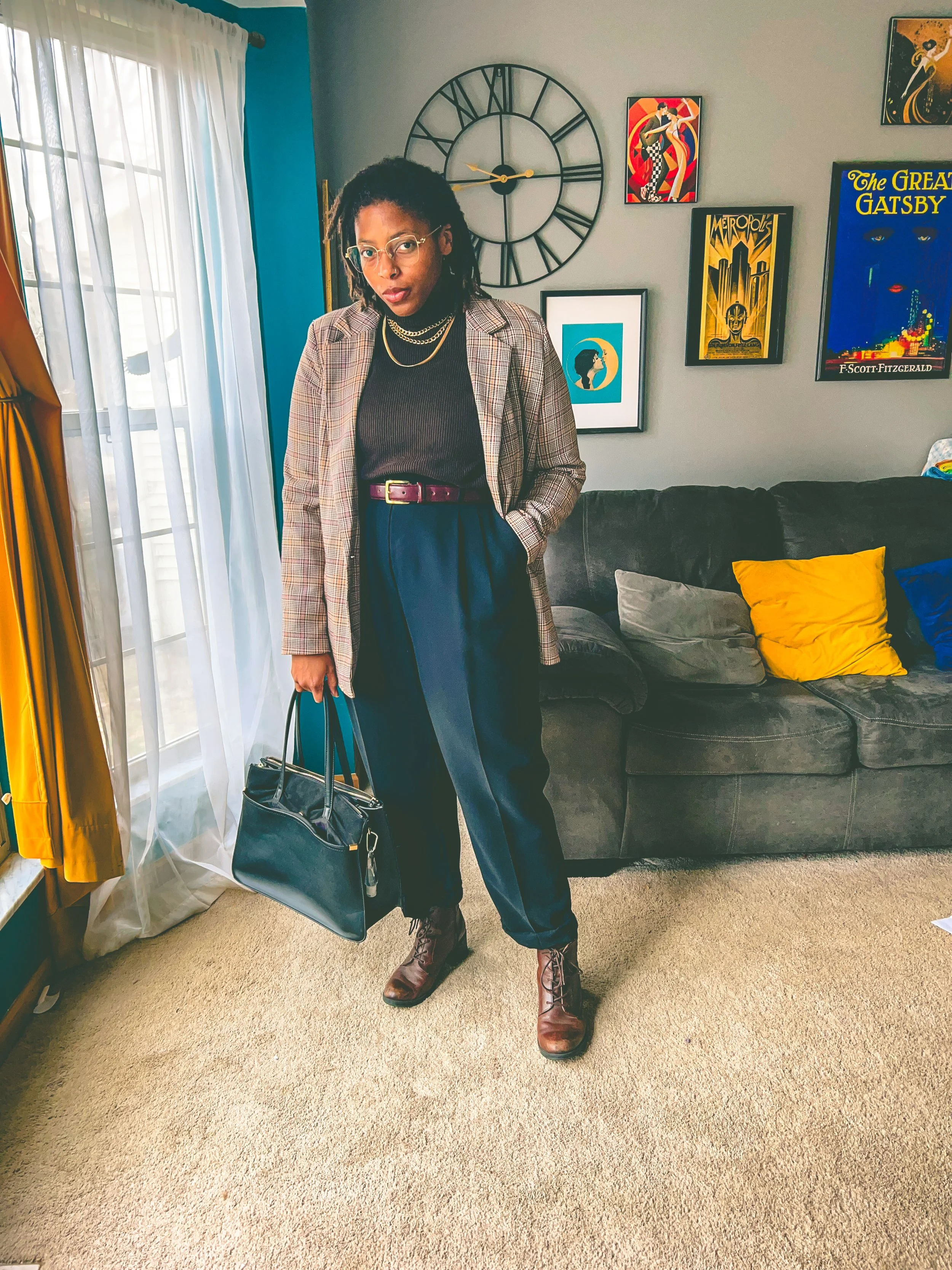 A woman standing in a living room, holding a black handbag, dressed in a plaid blazer, black top, dark trousers, and brown combat boots, with glasses and layered necklaces.