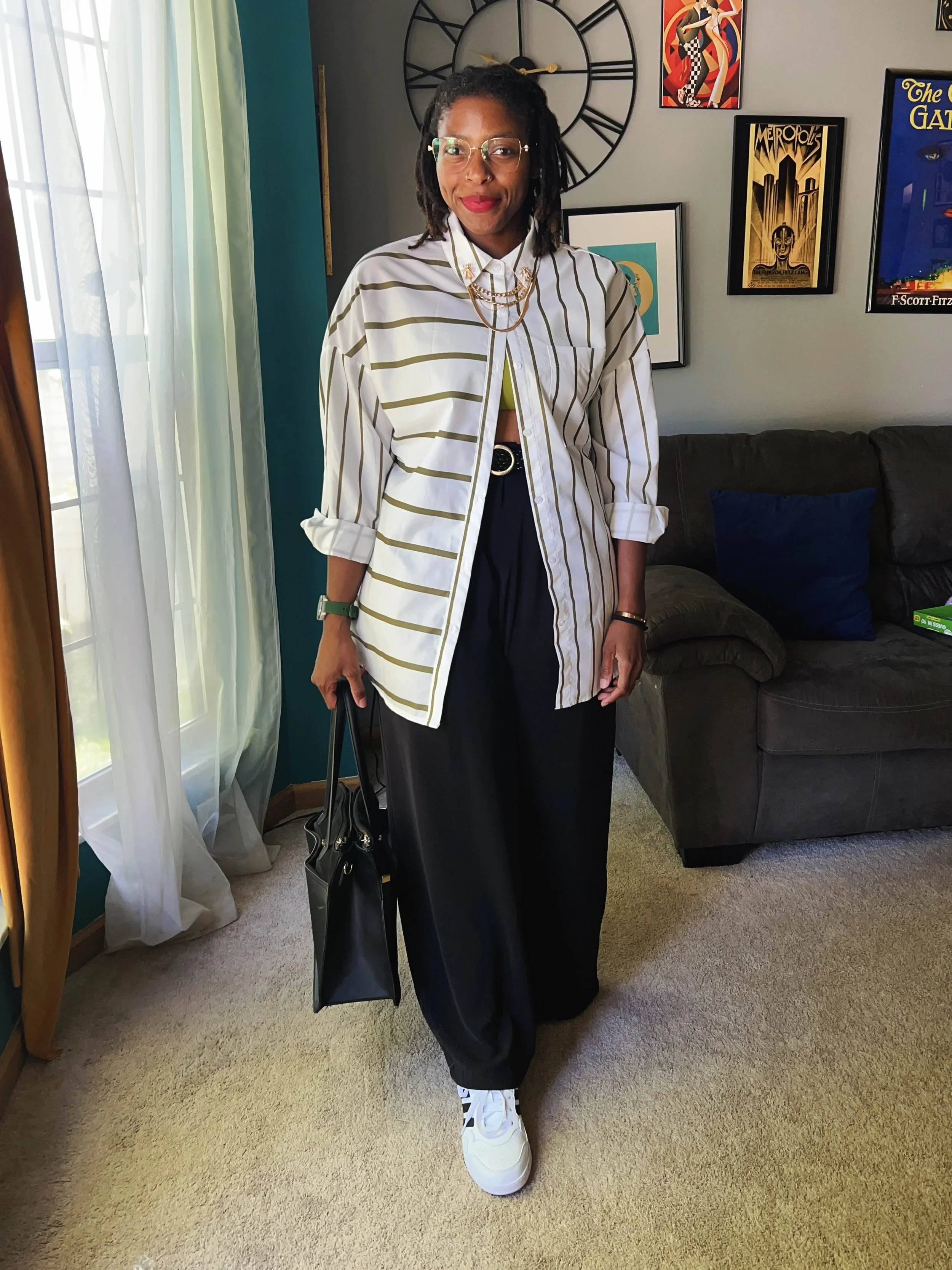 A woman with glasses, red lipstick, and dreadlocks standing in a living room, holding a black handbag. She wears a white and gold striped shirt over a neon green top, paired with black wide-leg pants and white sneakers. The room has a beige carpet, a