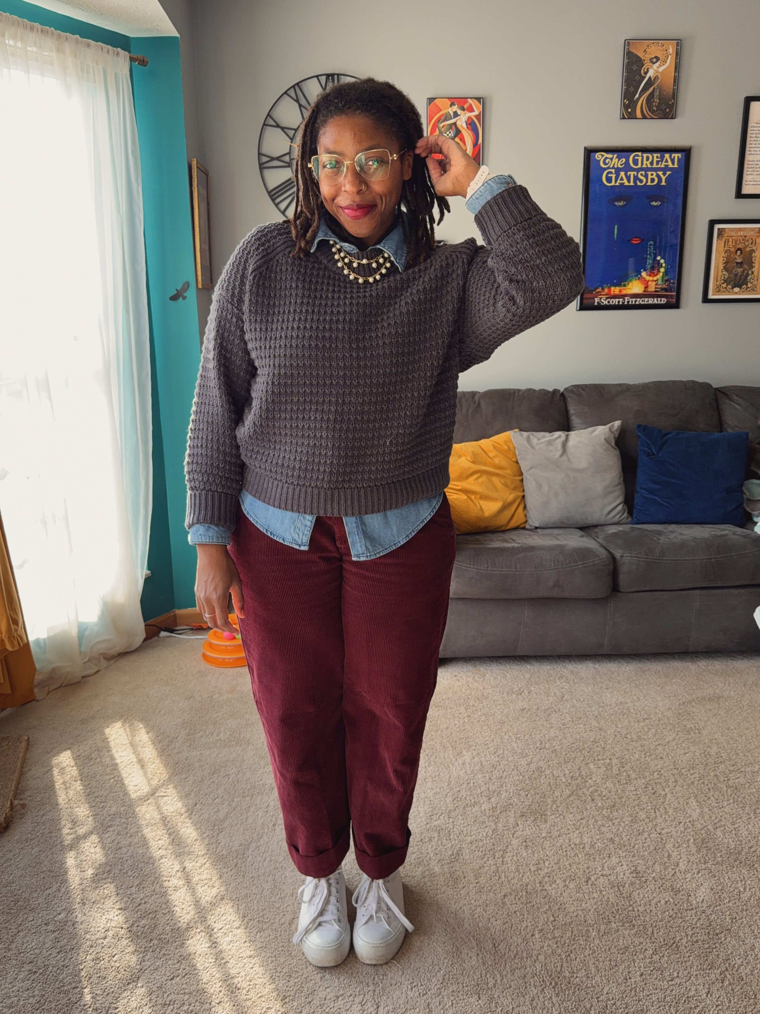 Woman with dreadlocks, glasses, red lipstick, wearing a gray sweater over a denim shirt, maroon pants, and white sneakers, standing in a living room.
