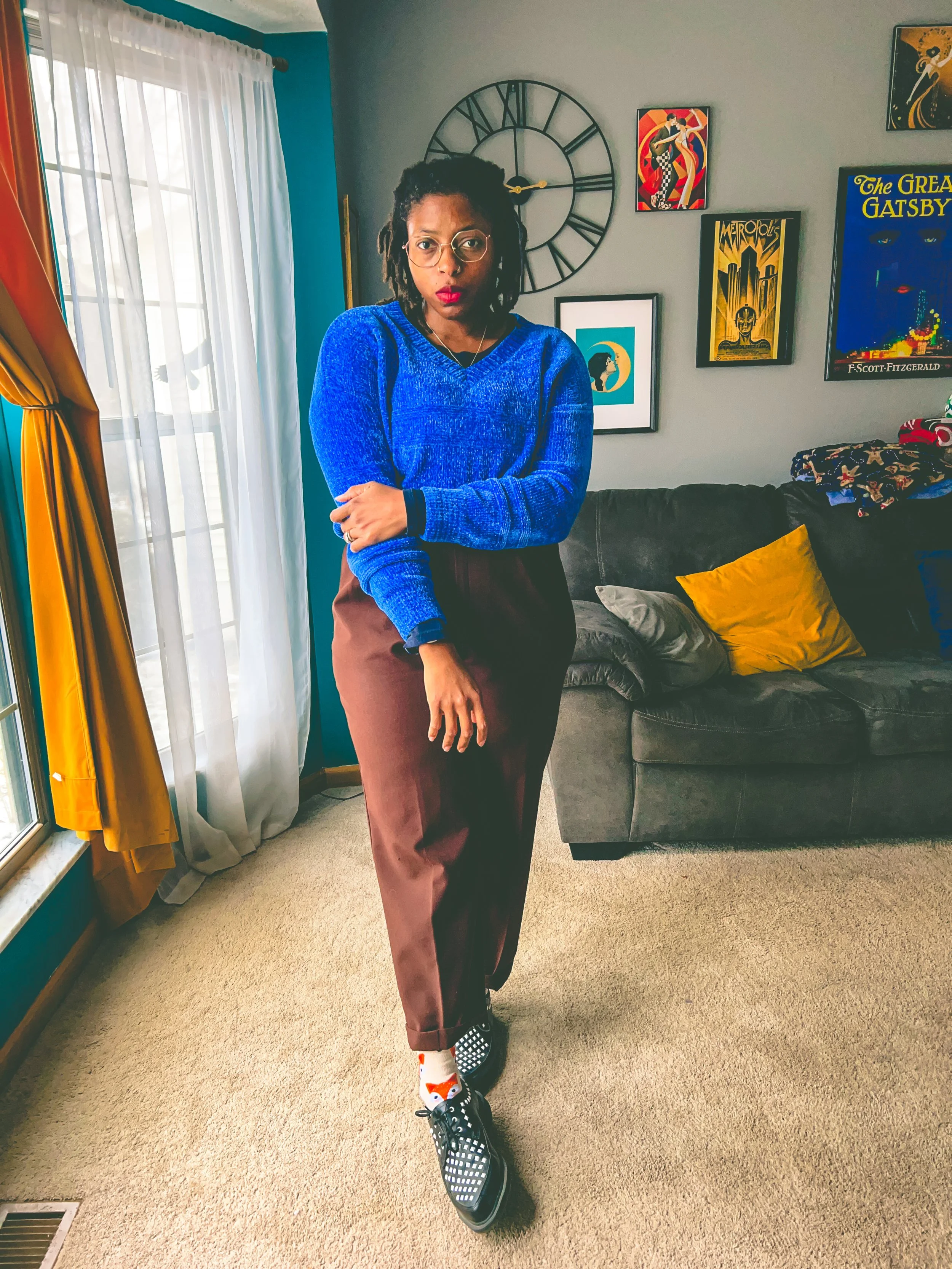 A woman with glasses and dreadlocks standing in a living room near sunlight, wearing a blue sweater, brown pants, colorful socks, and checkered shoes, with framed artwork and a large wall clock in the background.