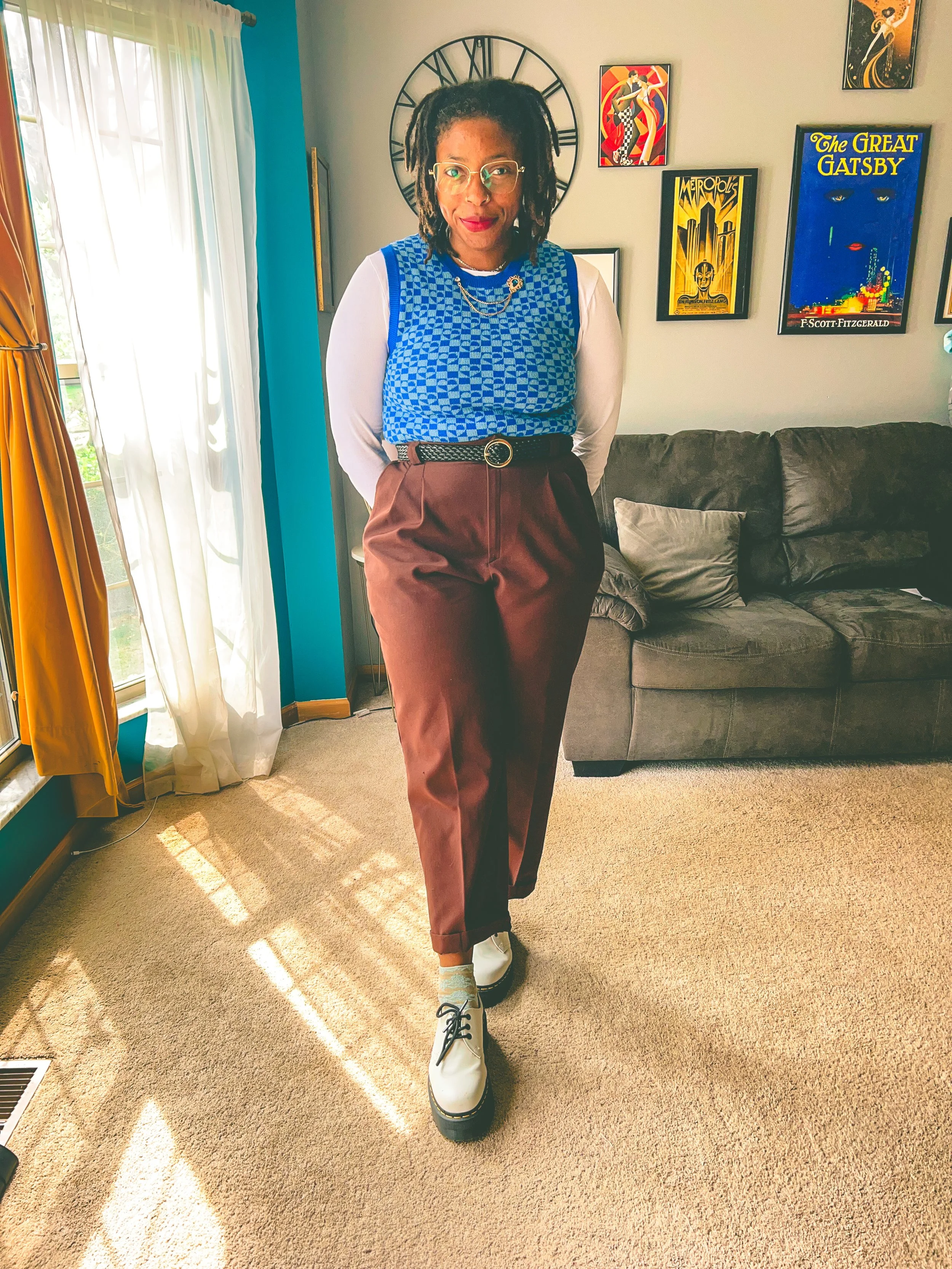 Woman standing indoors in front of a grey couch with a beige pillow, wearing glasses, a blue patterned top with white long-sleeve shirt underneath, brown pants, and white shoes, with framed artwork on the walls and a large clock behind her.
