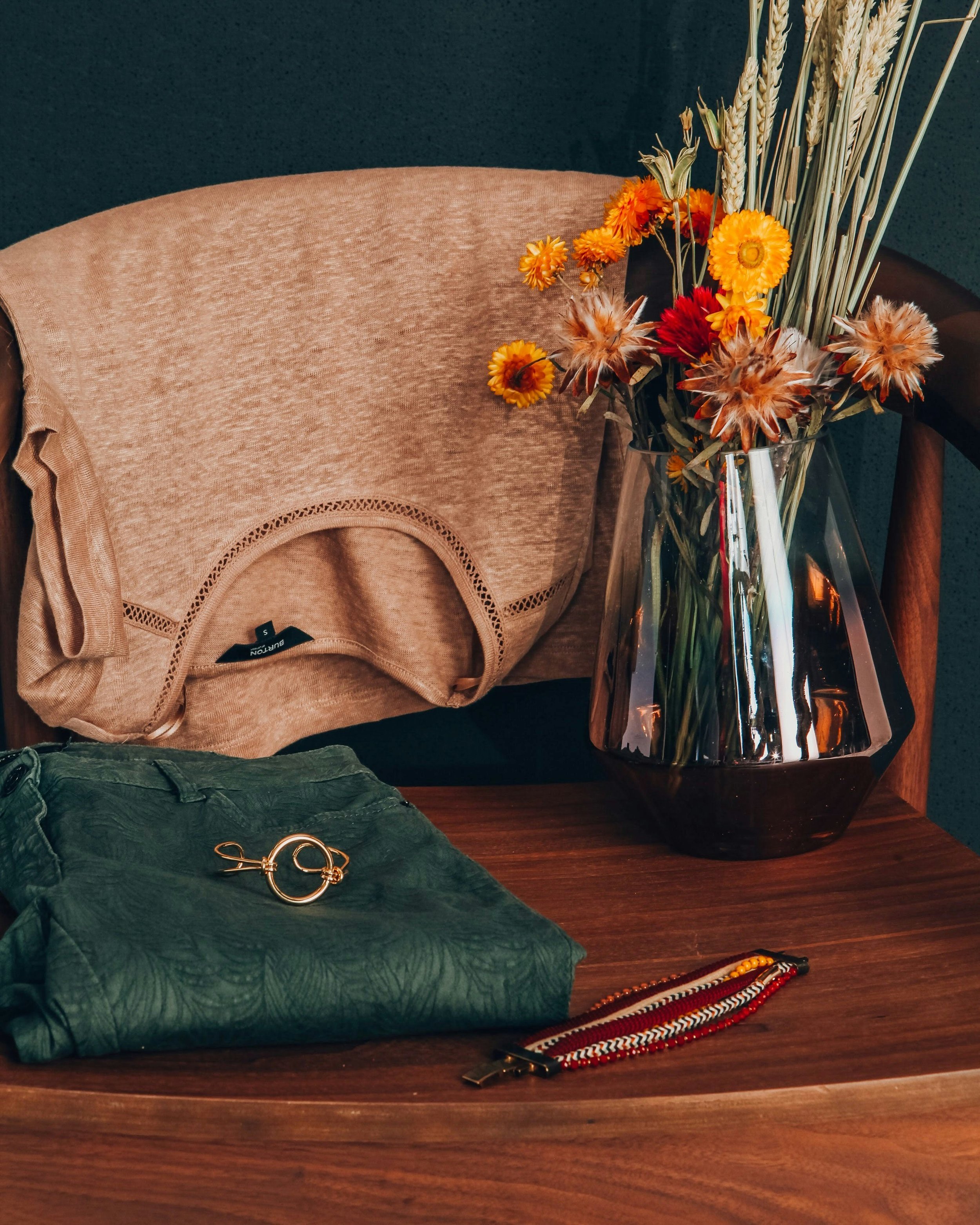 A beige Burts Bees shirt draped over a pink upholstered chair, a pair of green textured pants with a gold hair clip resting on top, a beaded bracelet, and a large glass vase with a colorful arrangement of yellow, orange, and brown dried flowers and grasses on a wooden table.