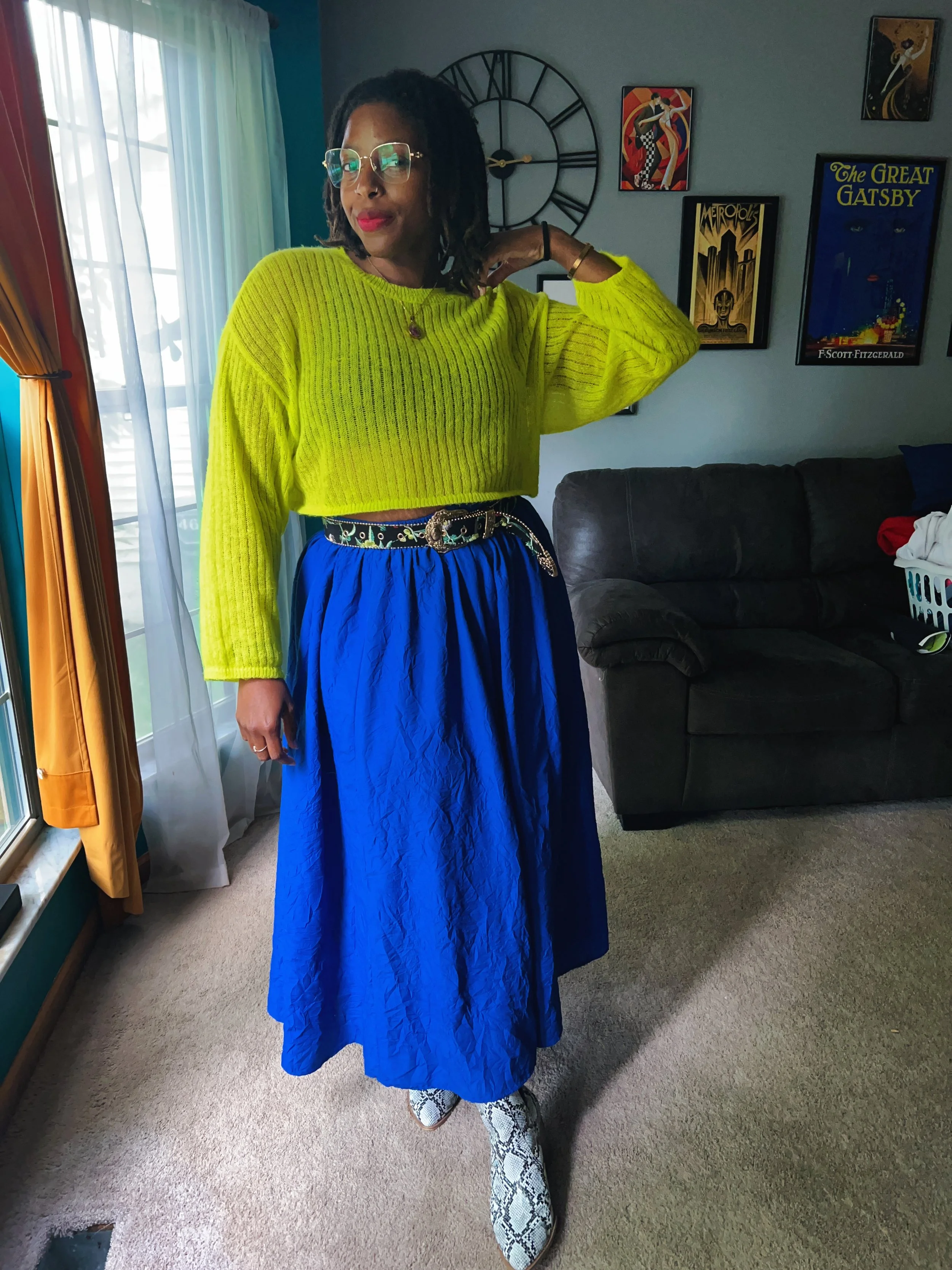 A woman wearing a bright yellow sweater, a long blue skirt, snakeskin boots, and glasses poses inside a living room with wall art and a large clock in the background.