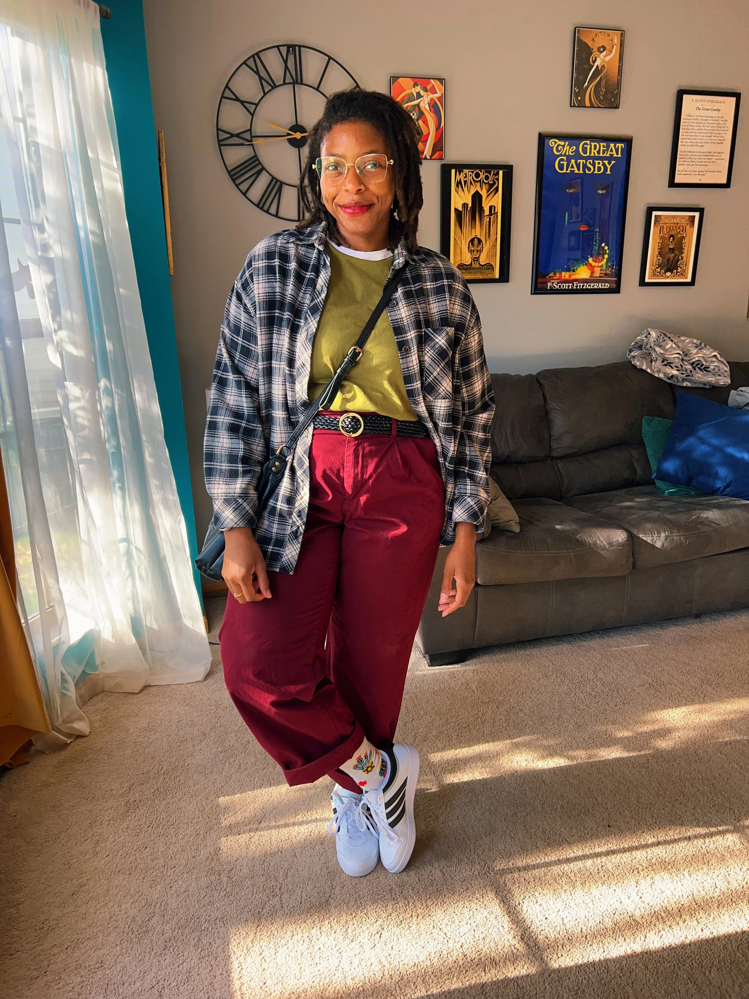 A woman with glasses and dreadlocks, dressed in a yellow shirt, plaid shirt, and red pants, standing in a living room with a large wall clock and framed artwork, smiling at the camera.