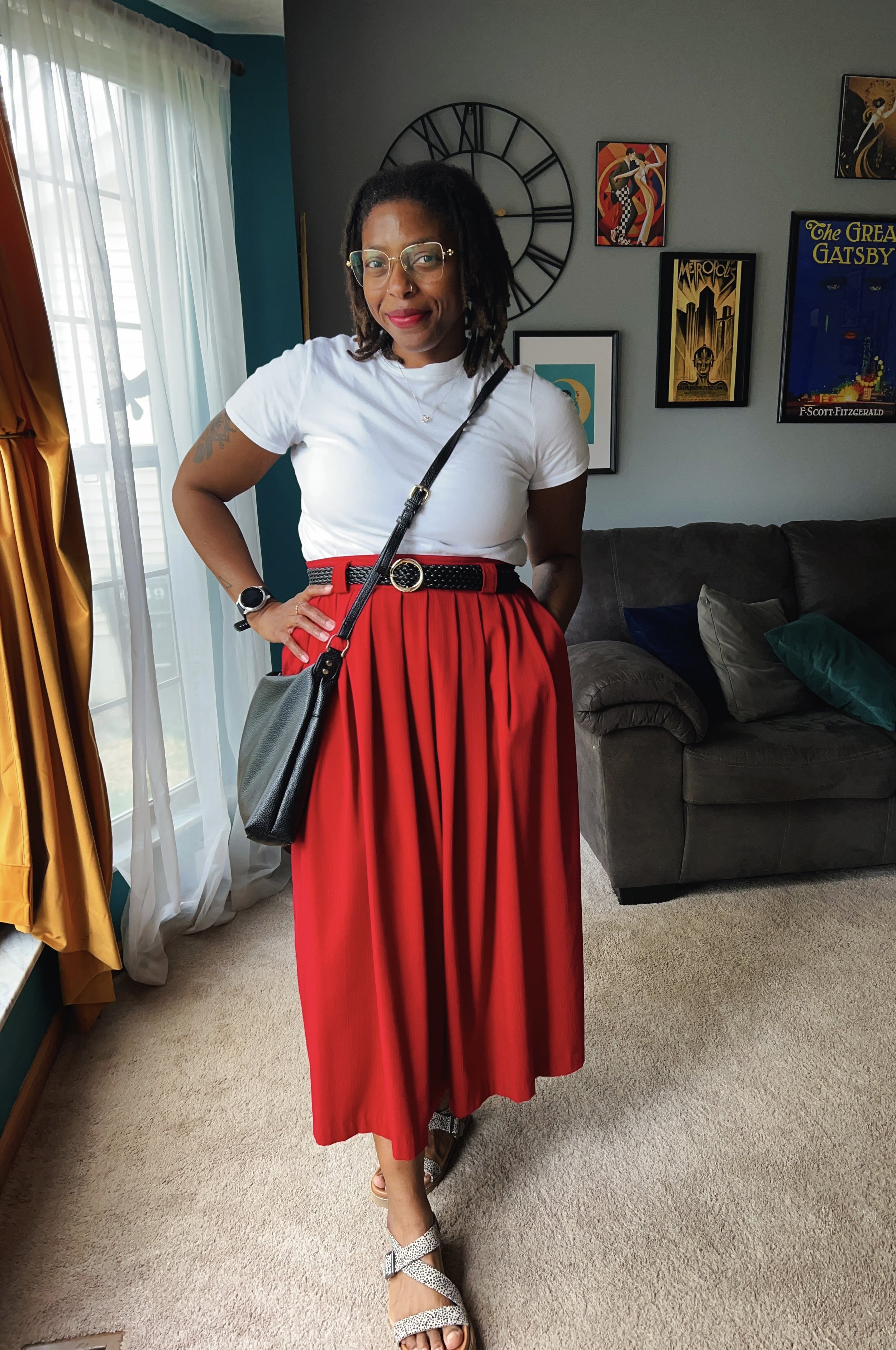 A woman standing indoors in front of a wall with artwork, wearing a white t-shirt, red pleated skirt, and sunglasses, carrying a black purse, and standing near a large window with white curtains.