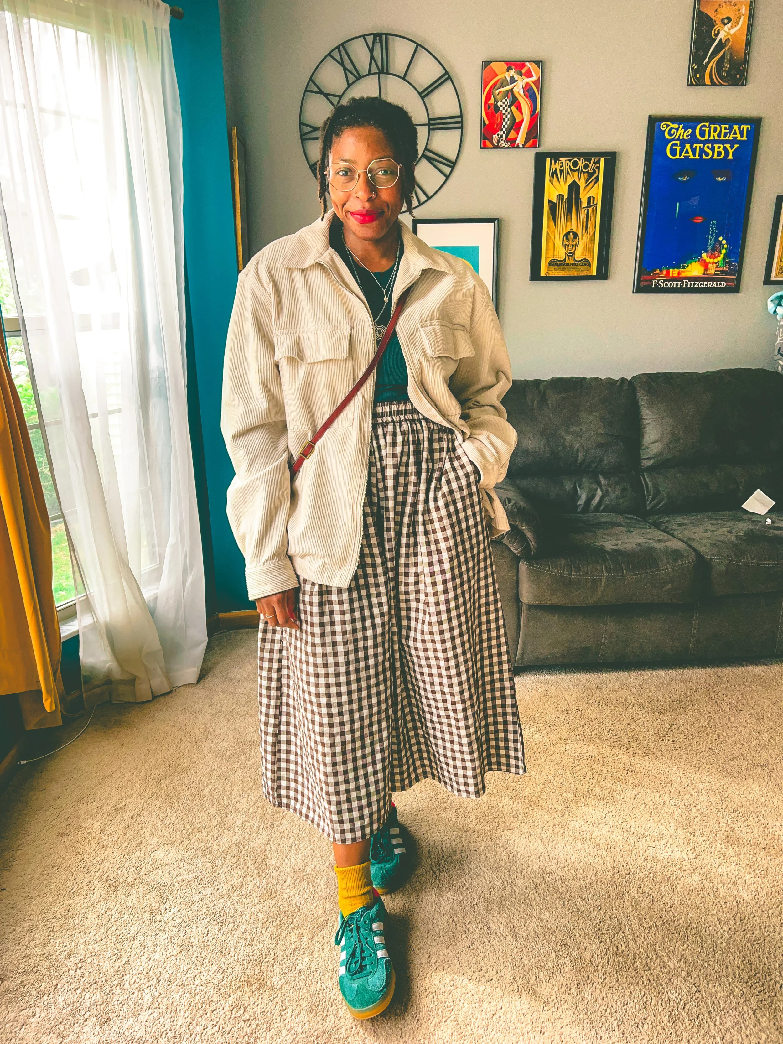 A woman with glasses, wearing a beige jacket, black top, and checkered wide-leg pants, standing in a living room with a black couch, colorful artwork on the wall, and curtains by a window.