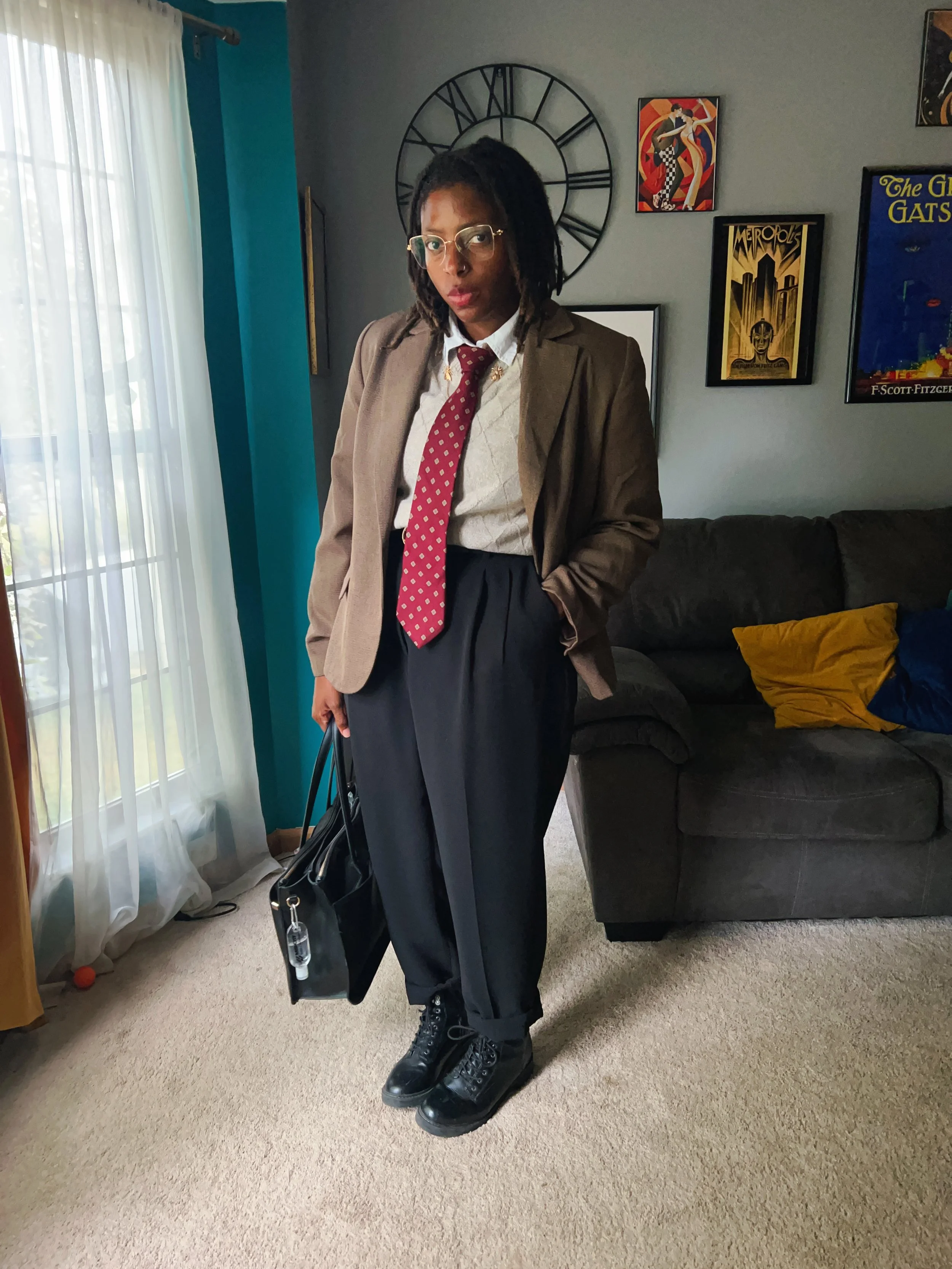 A woman standing in a living room, wearing glasses, a beige blazer, a white shirt, a red polka dot tie, black pants, and black combat boots, holding a black handbag.