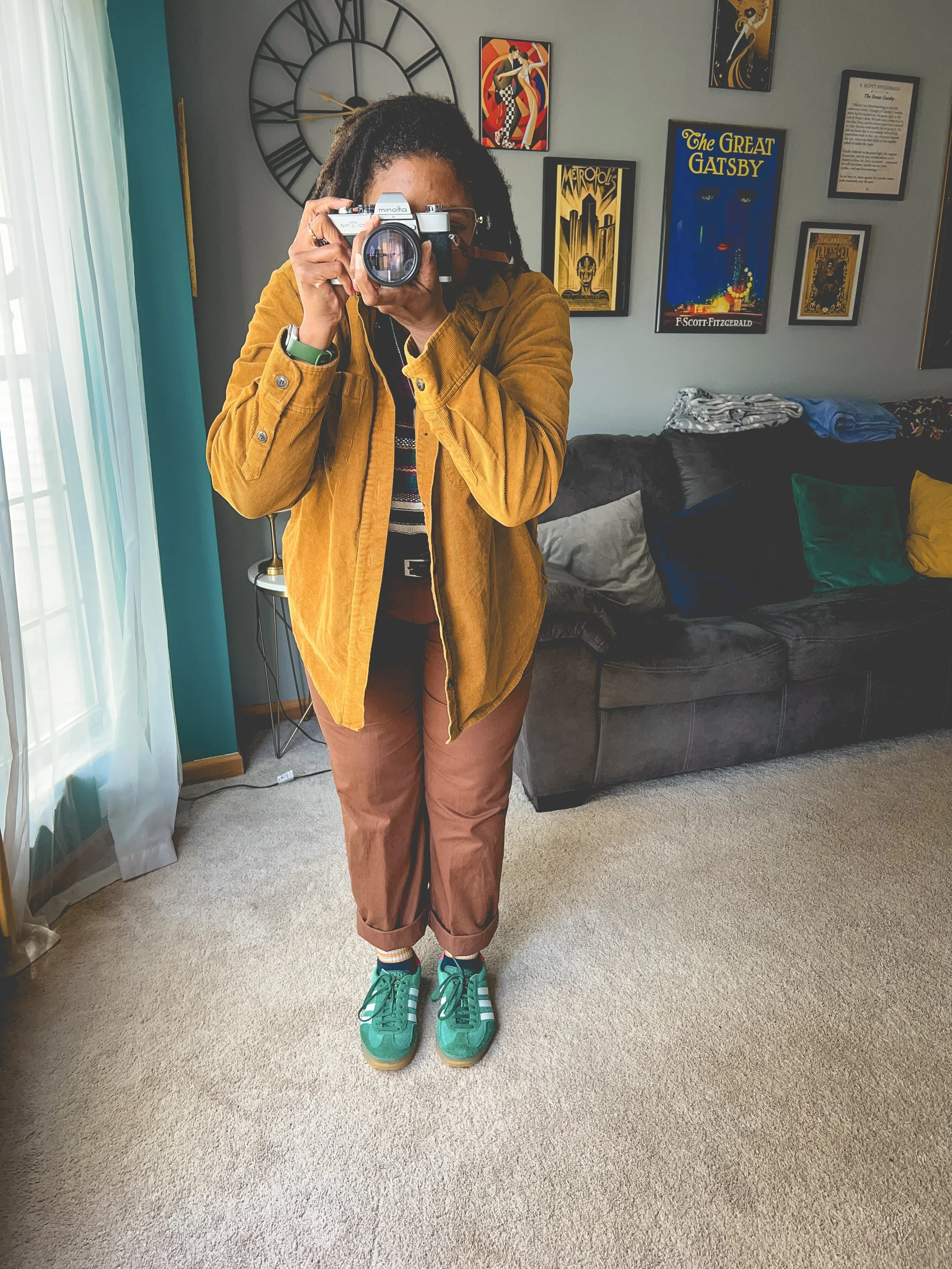 Person taking a mirror selfie with a camera in a living room, with a large wall clock and framed posters of 'Metropolis' and 'The Great Gatsby' on the wall behind them.