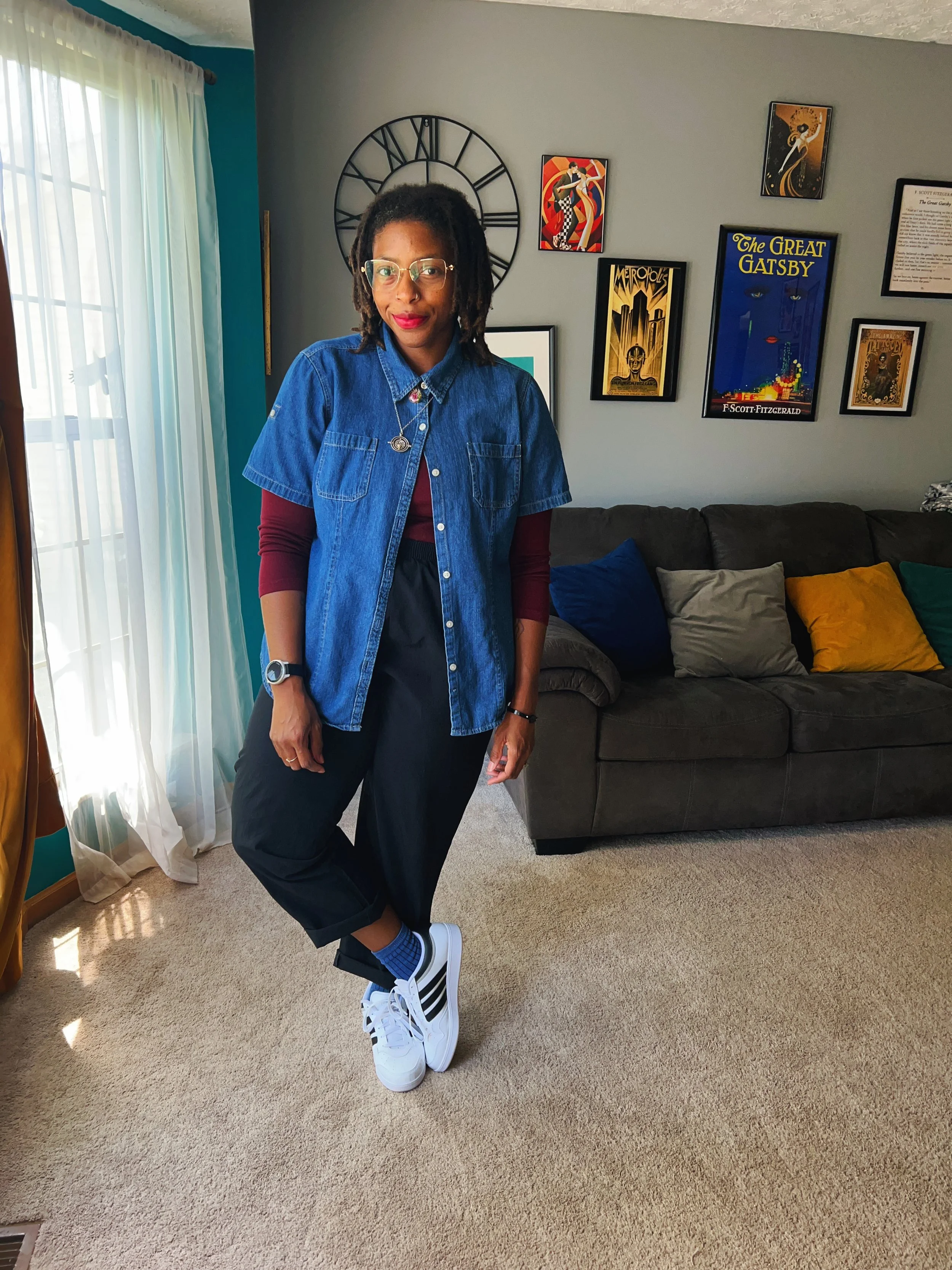 Woman standing in living room wearing glasses, denim shirt over maroon long sleeve shirt, black pants, sneakers, and blue socks. She has dreadlocks and is smiling.