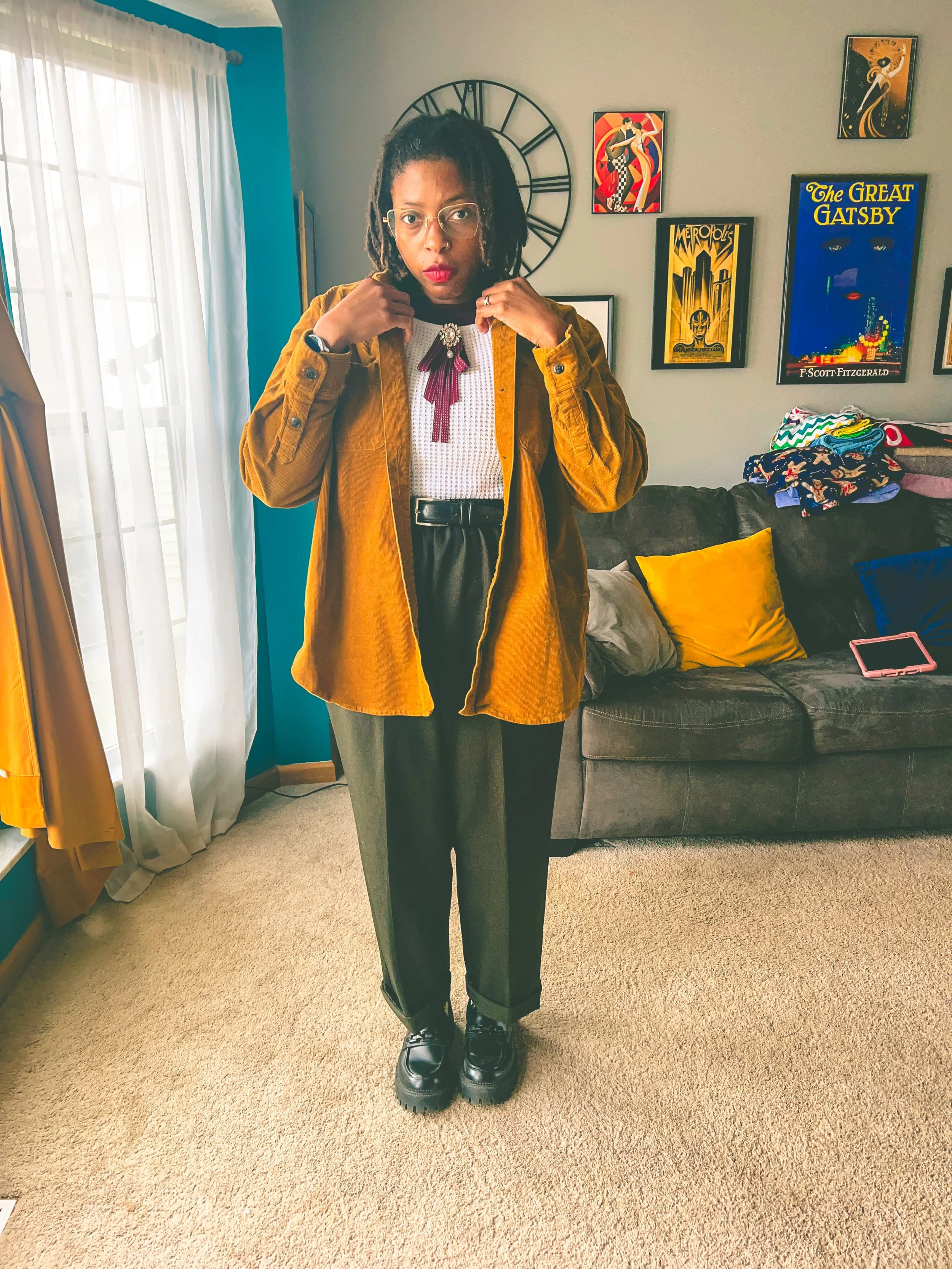 A woman standing in a living room, adjusting her necklace, wearing glasses, a mustard jacket, white top, and dark pants, with a sofa and colorful artwork in the background.