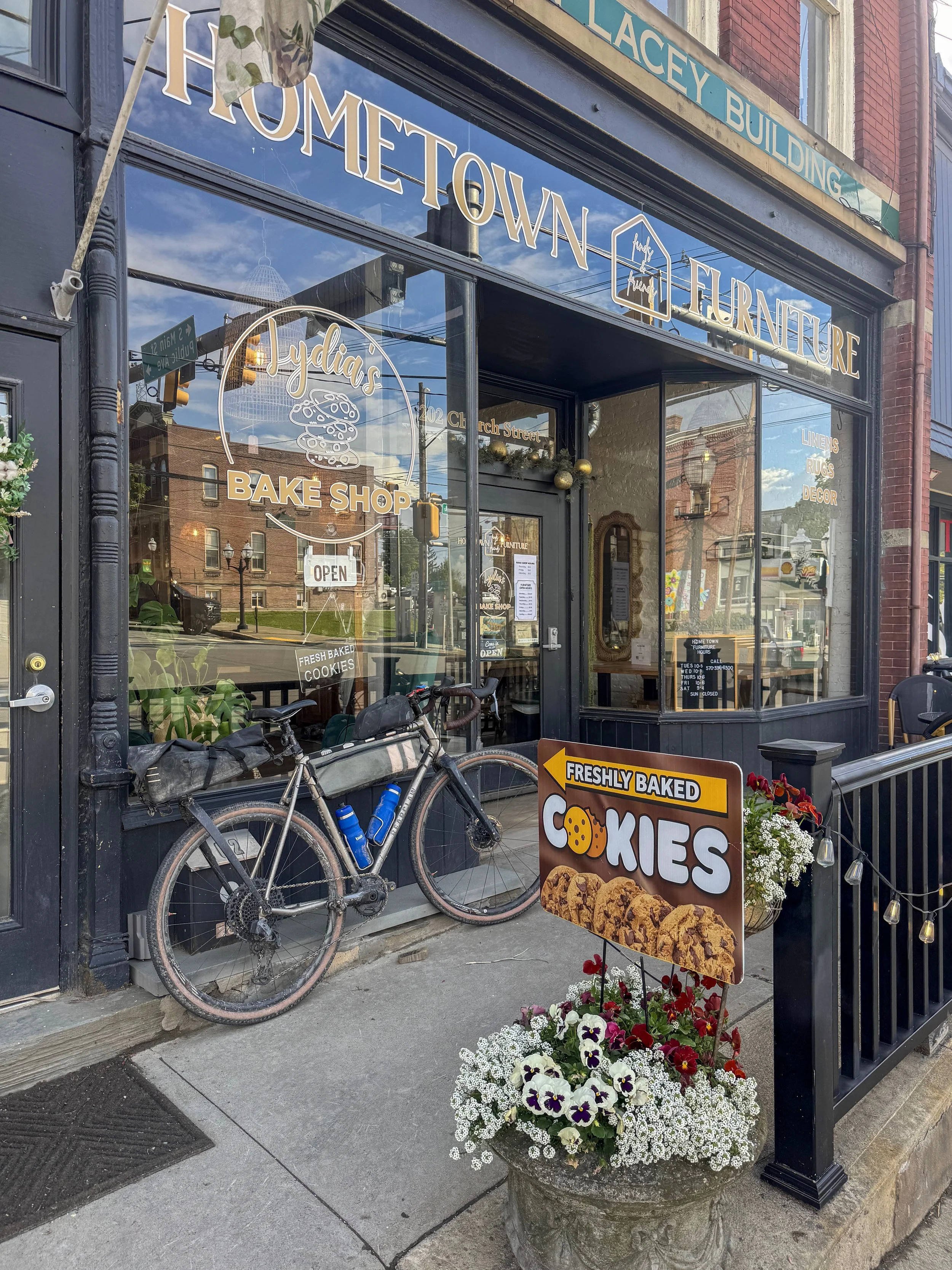 Exterior view of Lydia's Bake Shop, a bakery offering fresh baked cookies, with a bicycle parked outside, a colorful flower arrangement in a stone planter, a sign advertising cookies, and the storefront featuring large glass windows and signage.