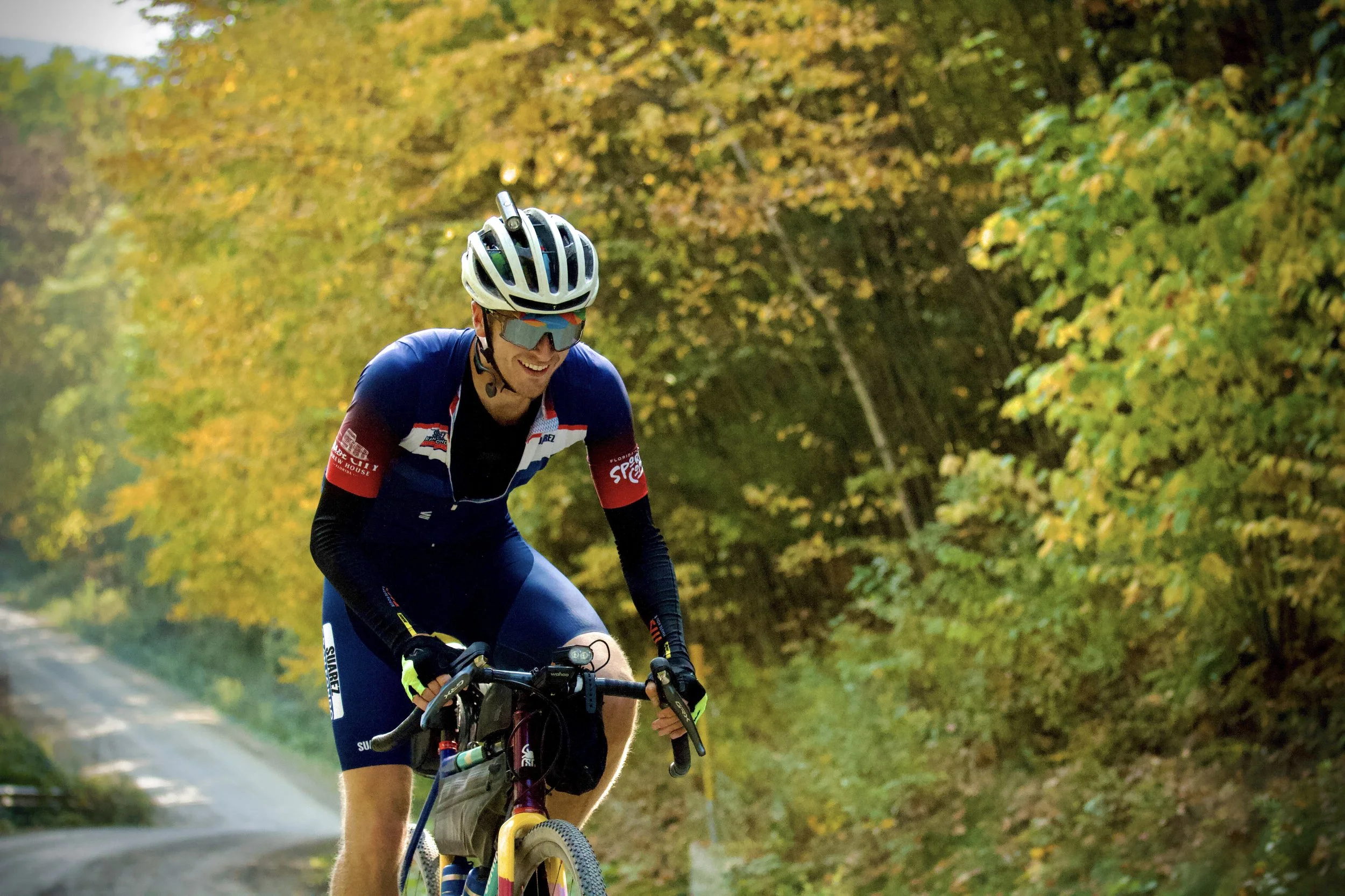 A person riding a bicycle on a tree-lined street during autumn, wearing a helmet, sunglasses, and a cycling jersey, smiling and enjoying the ride.