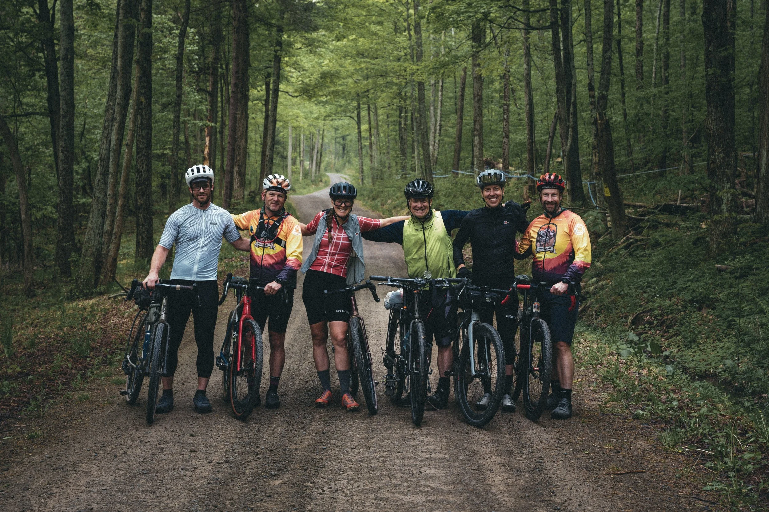 Group of six cyclists in helmets and athletic clothing, standing with bicycles on a forest trail, smiling and posing for the photo.