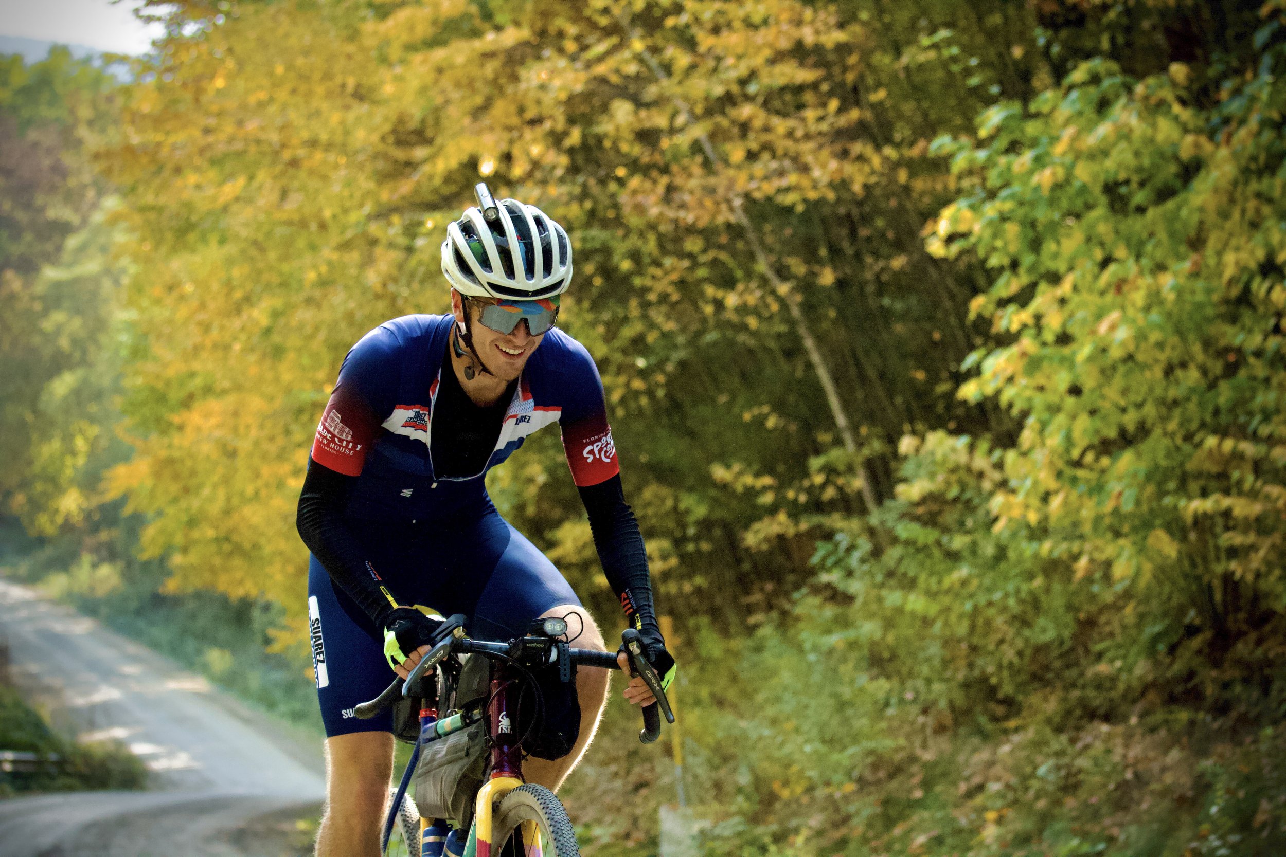 A man riding a mountain bike on a trail surrounded by autumn trees with yellow and orange leaves. He is wearing a helmet, sunglasses, and a blue and red cycling outfit, smiling as he pedals.