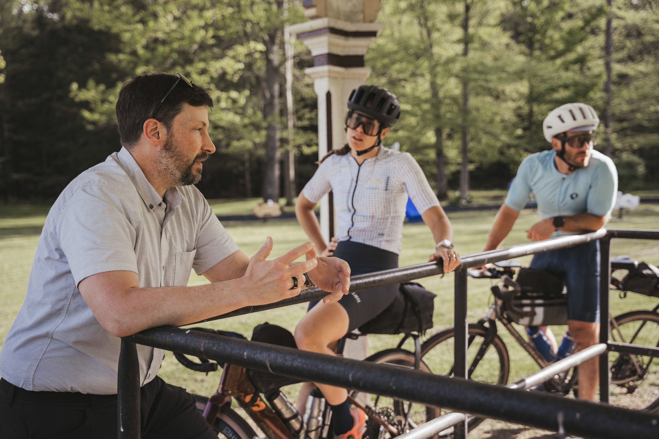 Three people, two men and one woman, are having a conversation outdoors near a park bench with bicycles. The man on the left, with a beard and wearing a light-colored shirt, is talking to the woman and man, who are both wearing helmets and sunglasses