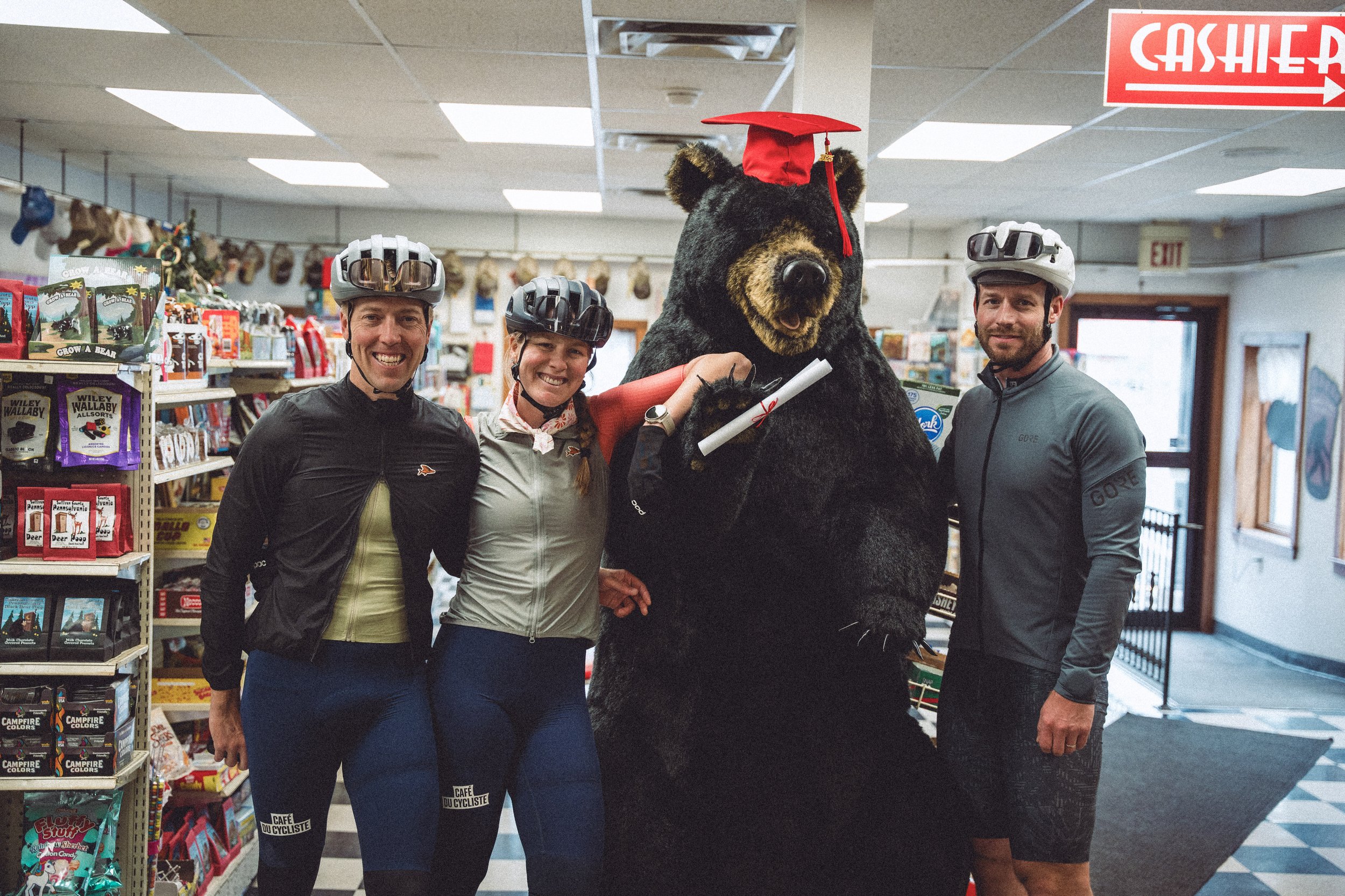 Four people standing in a store with a bear mascot wearing a red graduation cap. The group includes three cyclists wearing helmets and athletic clothing, smiling at the camera. The store shelves behind them have bags of candy and other items, and the