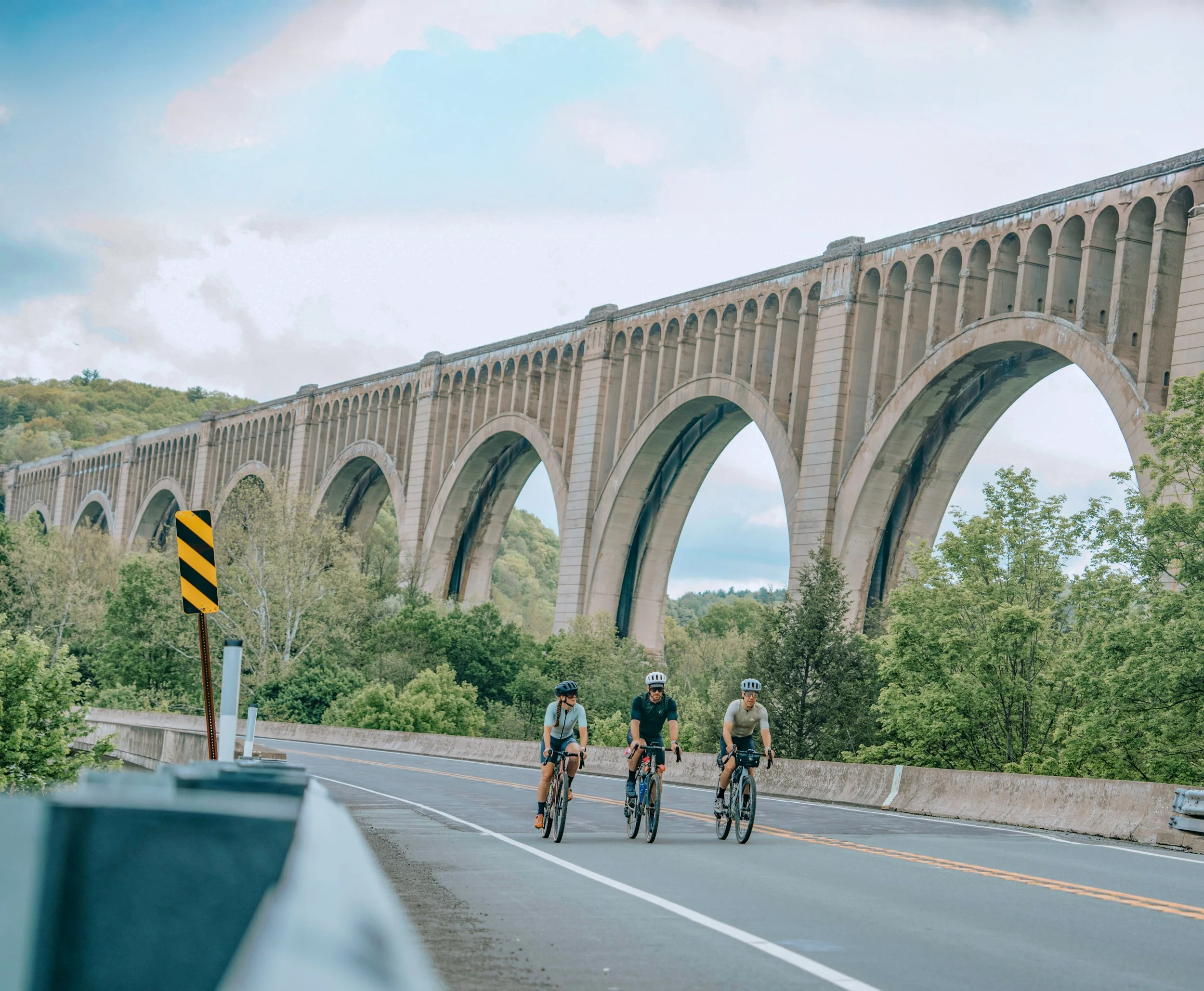 Four cyclists riding on a road beneath a large stone bridge with multiple arches, surrounded by green trees and hills, under a partly cloudy sky.