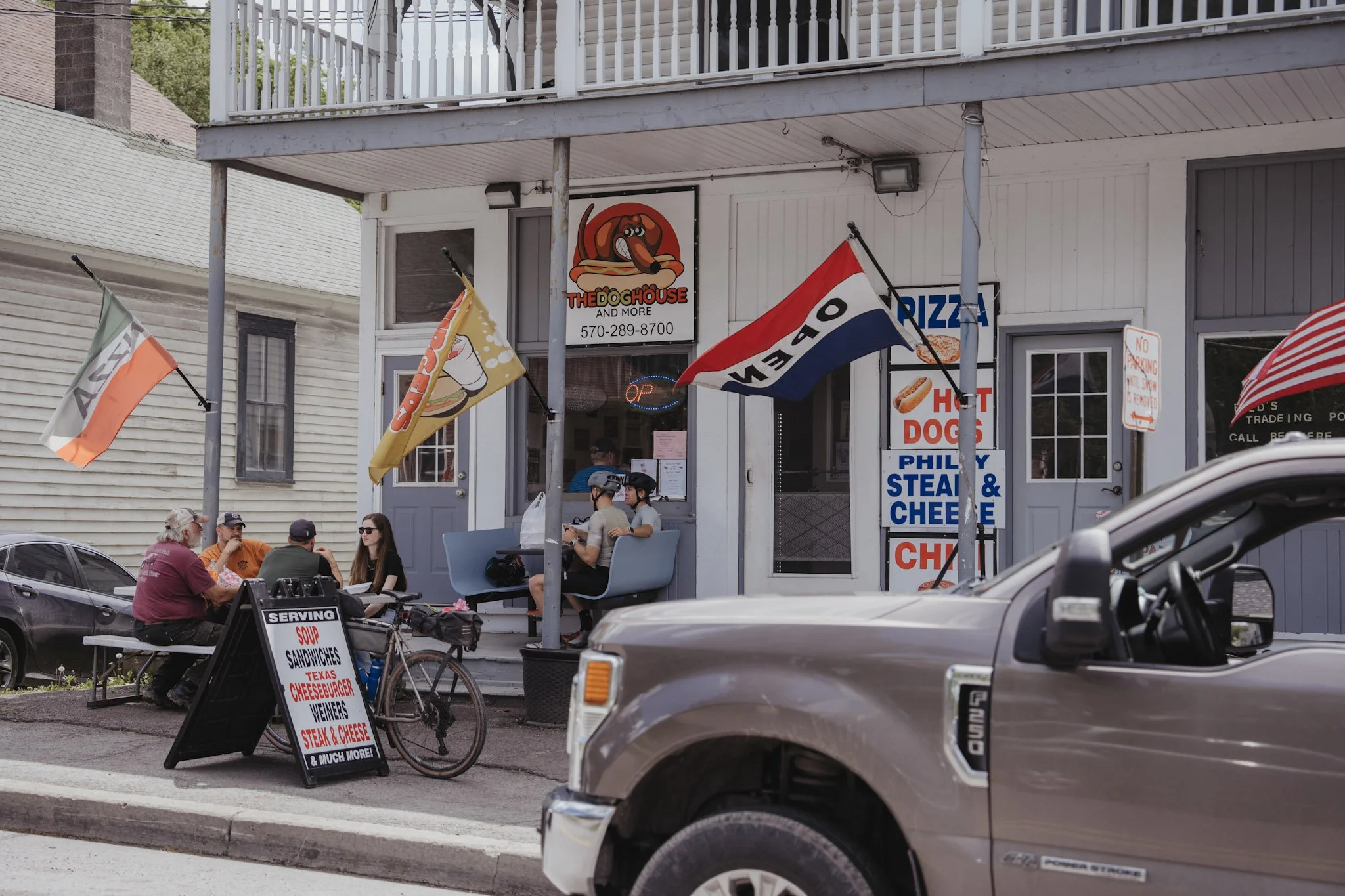 People sitting outside a restaurant called 'The Dog House,' with flags and signs on the sidewalk advertising hot dogs, sandwiches, and other fast food. The restaurant has a white exterior with a sign of a cartoon dog.