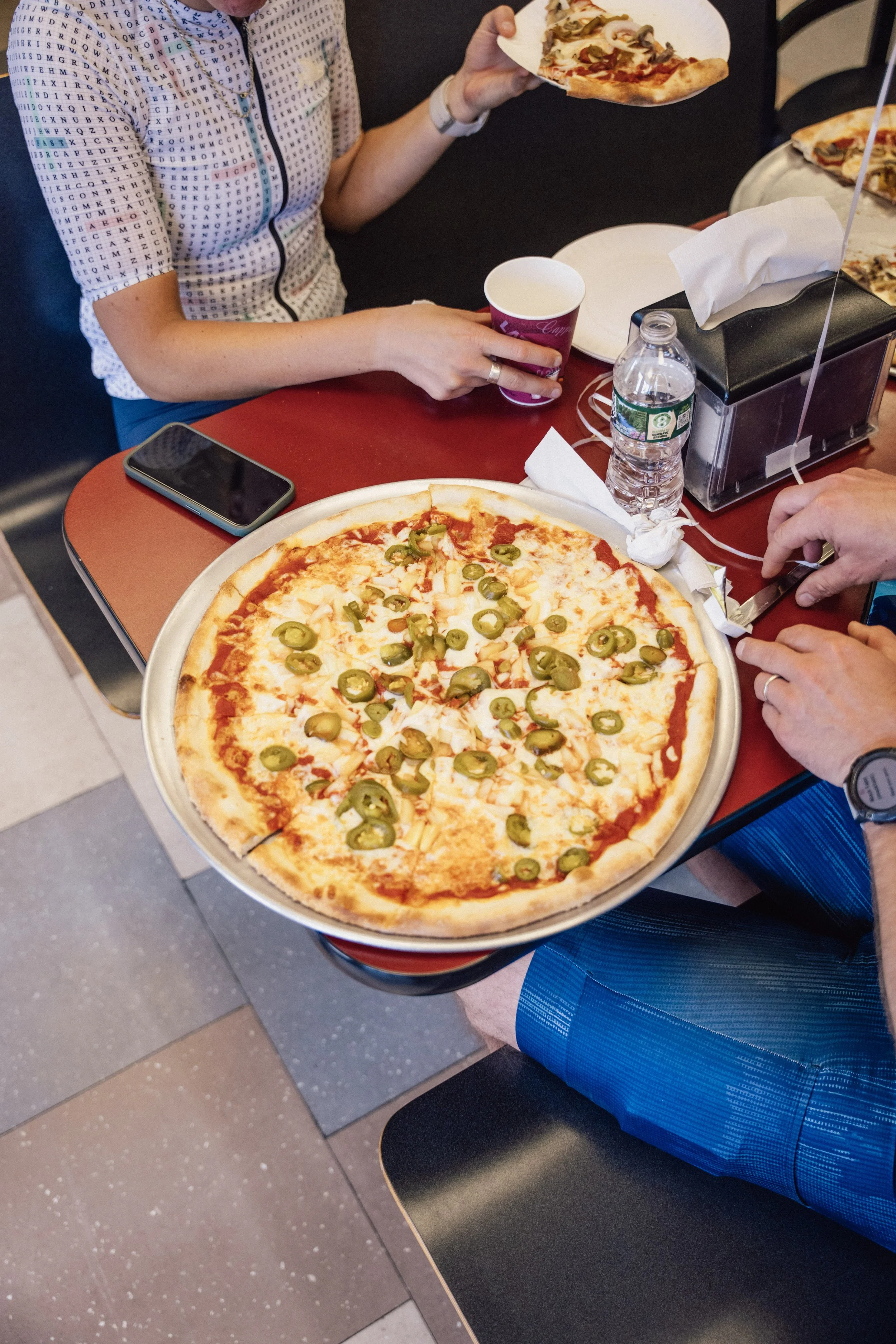 A person holding a slice of pizza with jalapeño peppers, cheese, and tomato sauce, seated at a table with additional slices and a whole pizza. The person is wearing a patterned shirt, and there are items on the table including a smartphone, a water b