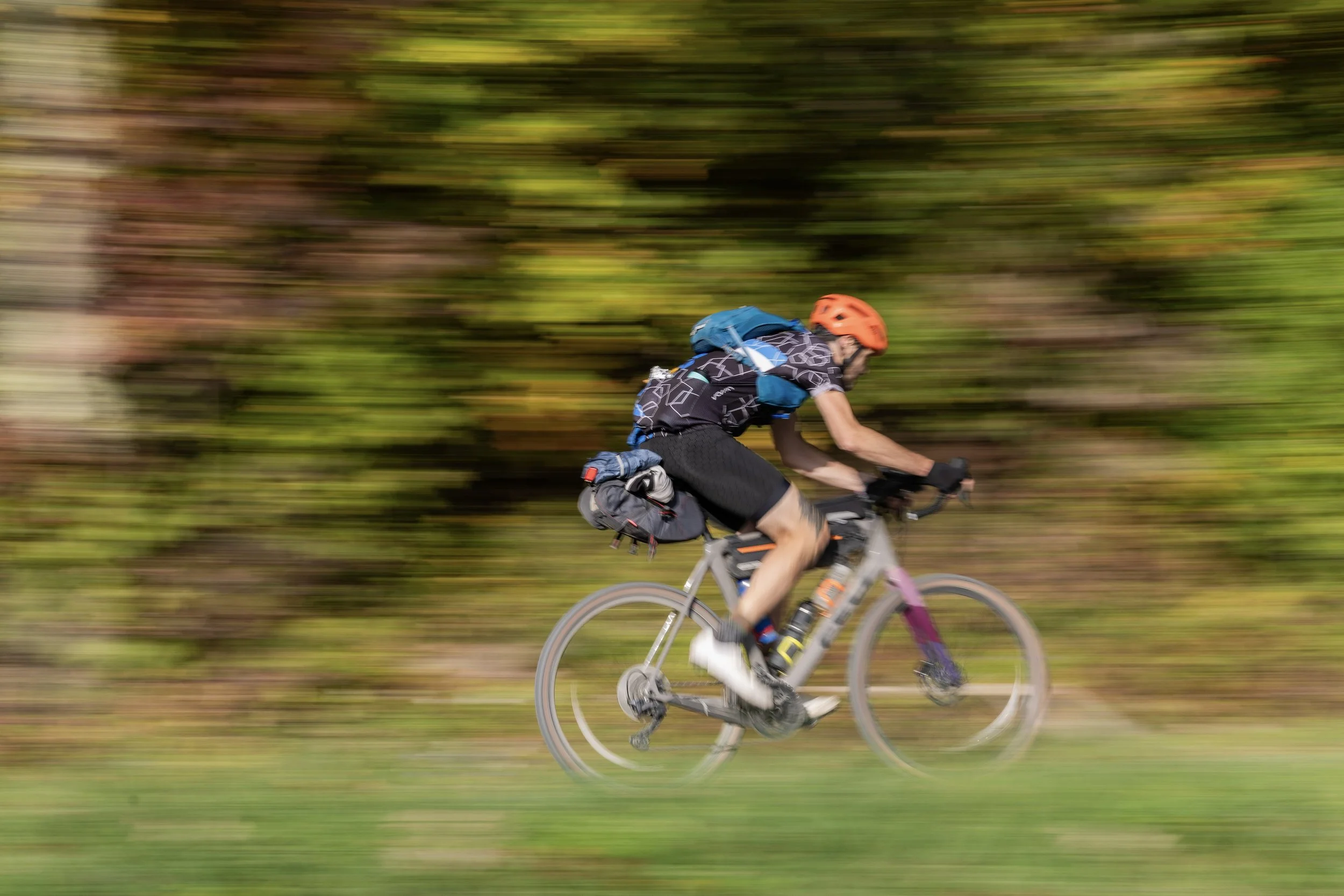 A person riding a mountain bike quickly through a forested trail, wearing an orange helmet, black cycling attire, and a backpack, with a motion blur background of trees and greenery.