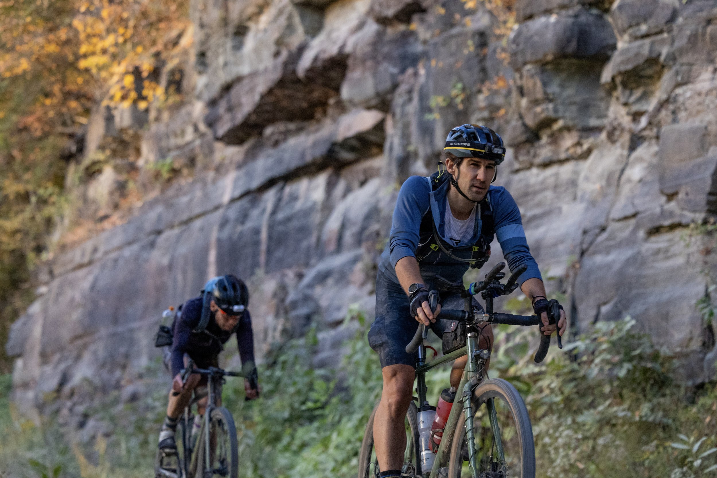 Two mountain bikers riding along a rocky trail in a forest with fall foliage, wearing helmets and biking gear.