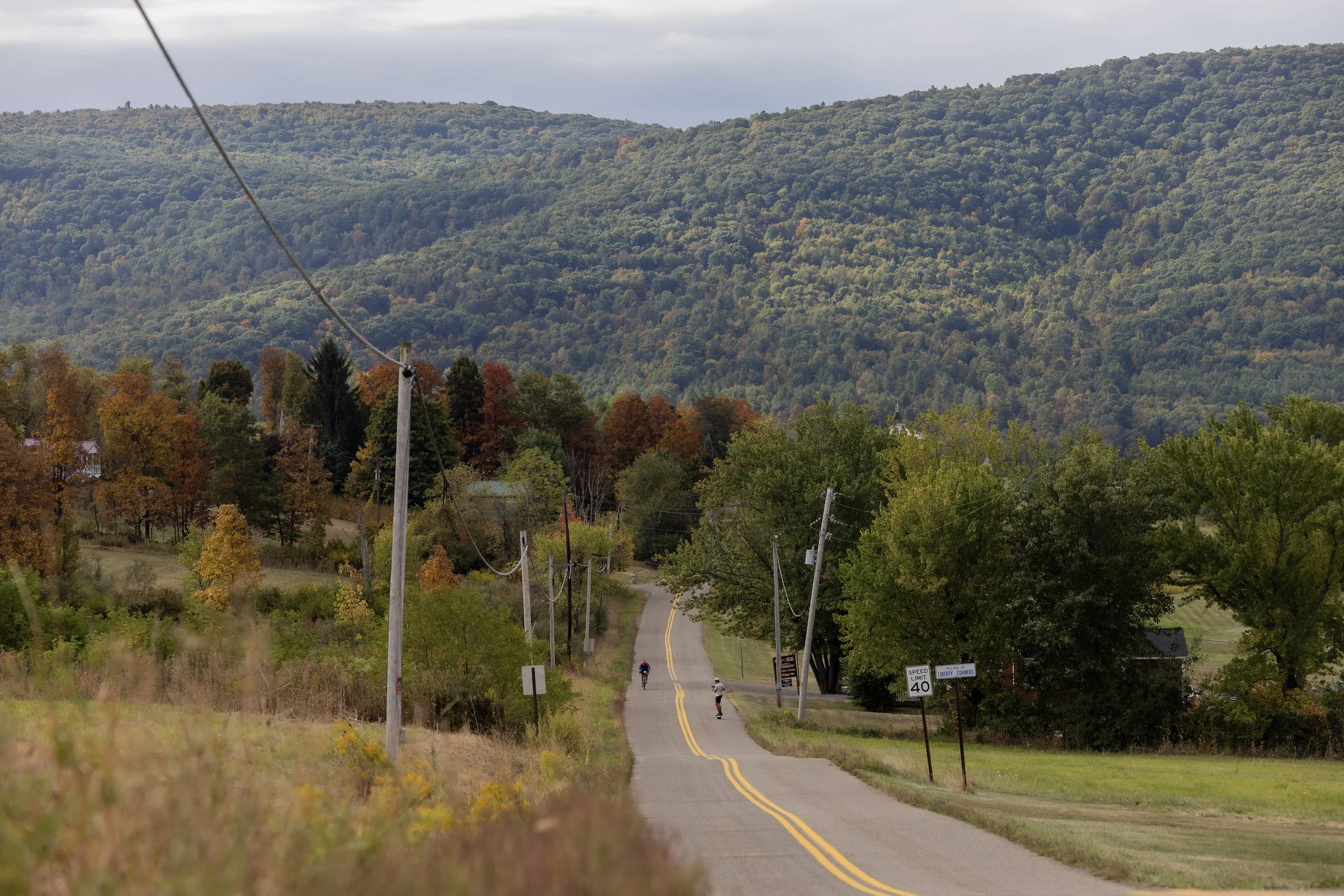 A rural road descending into a valley surrounded by tree-covered hills. A few cyclists are riding on the road, and there are utility poles along the roadside. The trees display a mix of green and autumn colors, and the sky is overcast.