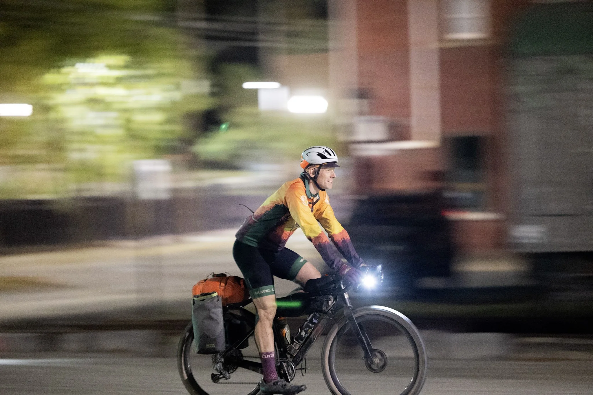 A cyclist wearing a helmet and colorful cycling gear riding a bicycle at night with blurred city lights and buildings in the background.
