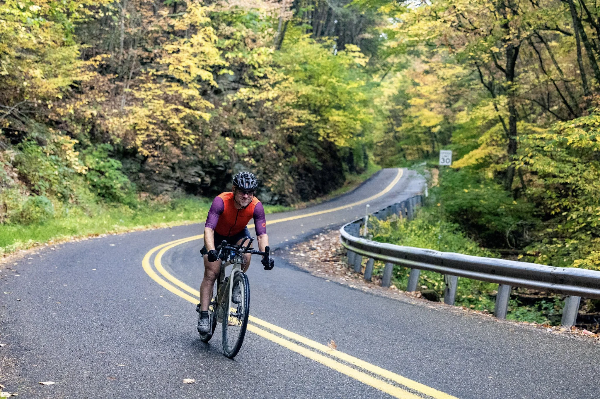 A man riding a bicycle along a winding road surrounded by trees with yellow and green leaves during fall.