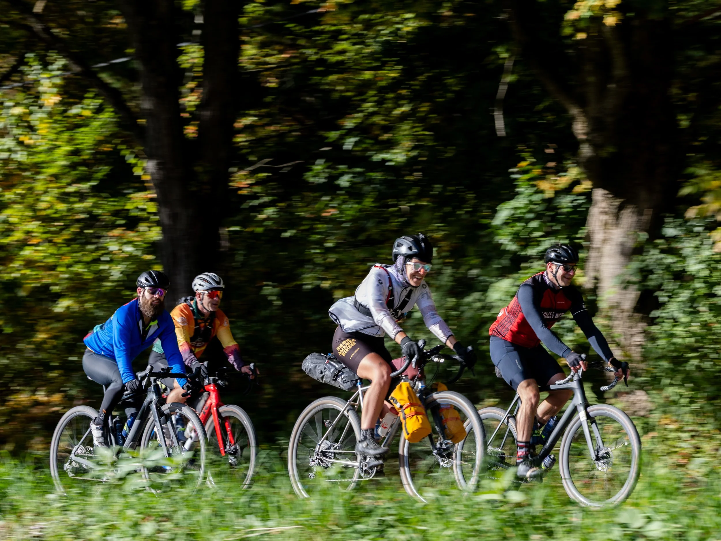 A group of people riding bicycles through a wooded area during the day, wearing helmets and outdoor clothing, smiling and enjoying the ride.