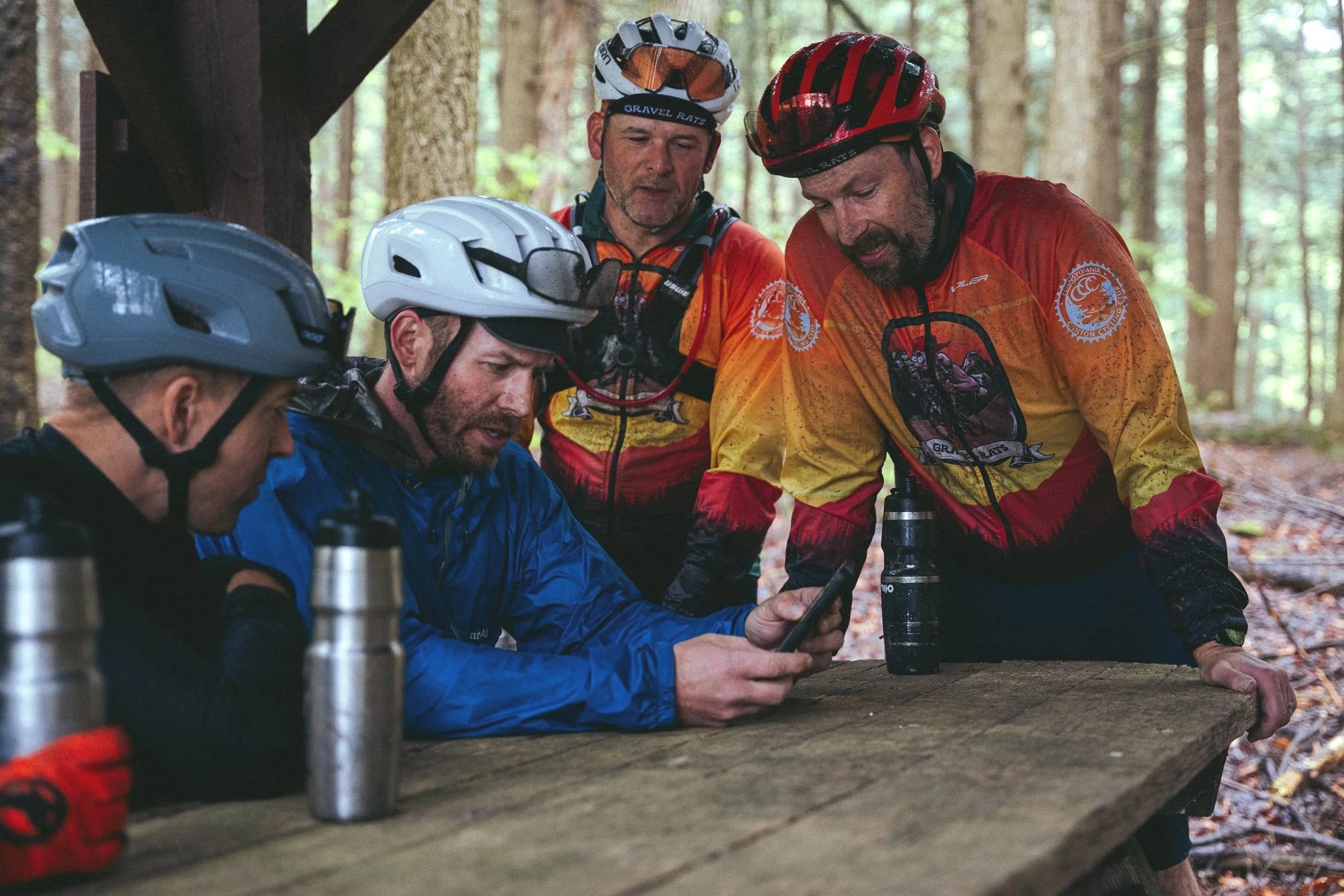 Four mountain bikers in helmets and colorful jerseys gather around a wooden picnic table in a forest, looking at a smartphone.
