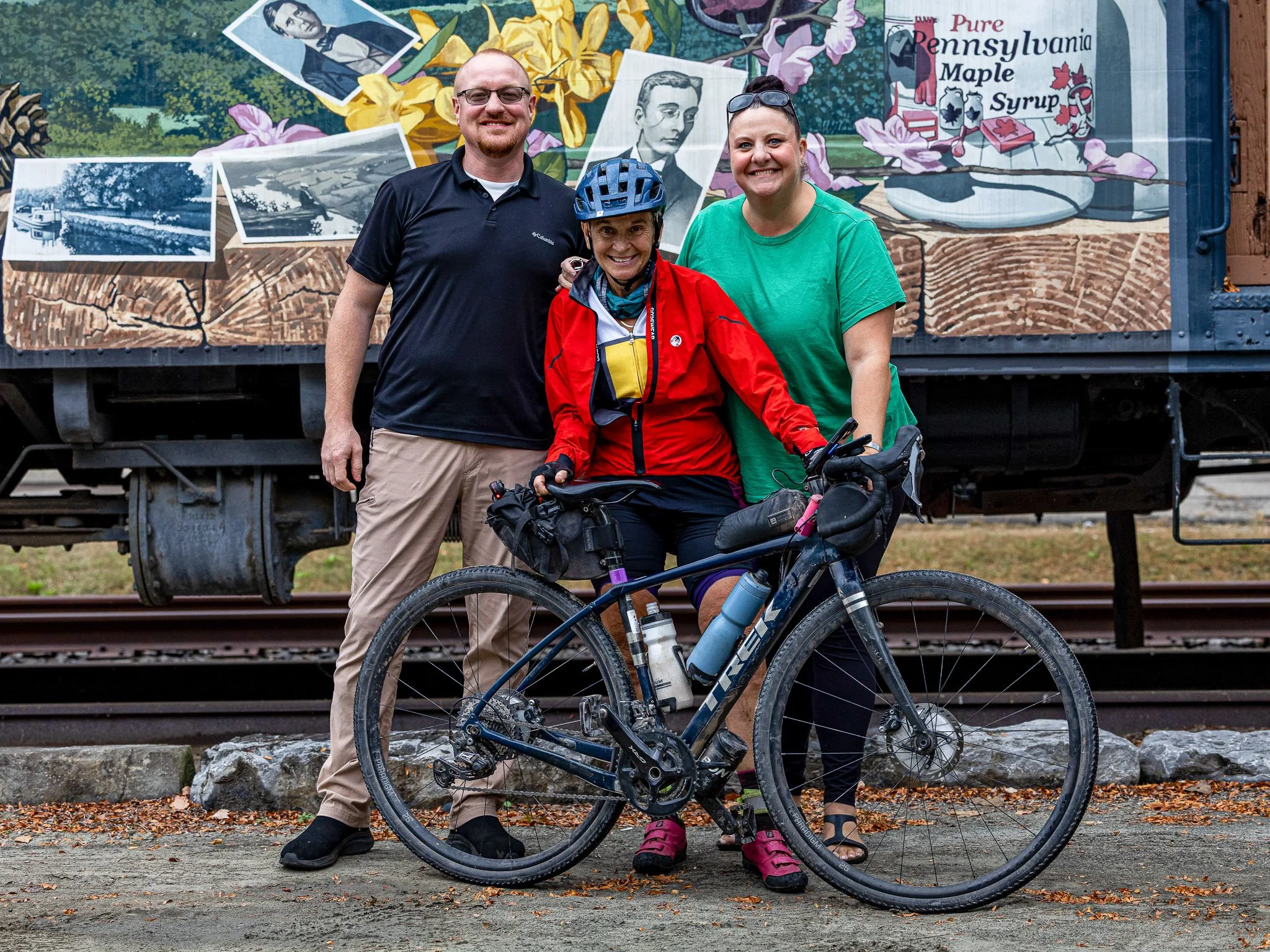 Three people standing outdoors in front of a colorful mural featuring historical photographs, flowers, and a sign advertising Pennsylvania maple syrup. One woman in cycling gear and a helmet is sitting on a bicycle, with two others standing beside her, smiling for the photo.