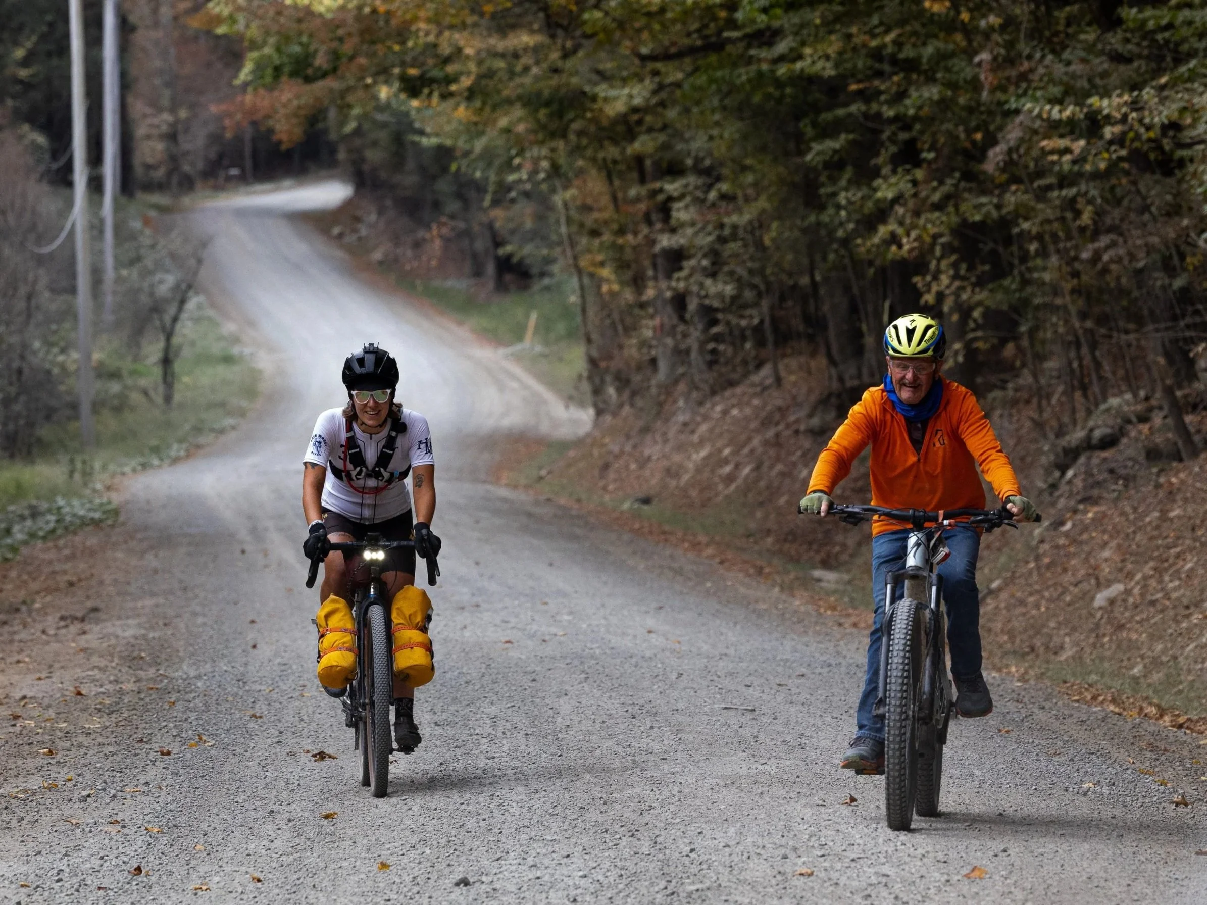 Two people riding mountain bikes on a dirt trail through a forest with fall foliage, one wearing a white shirt and the other wearing an orange jacket.