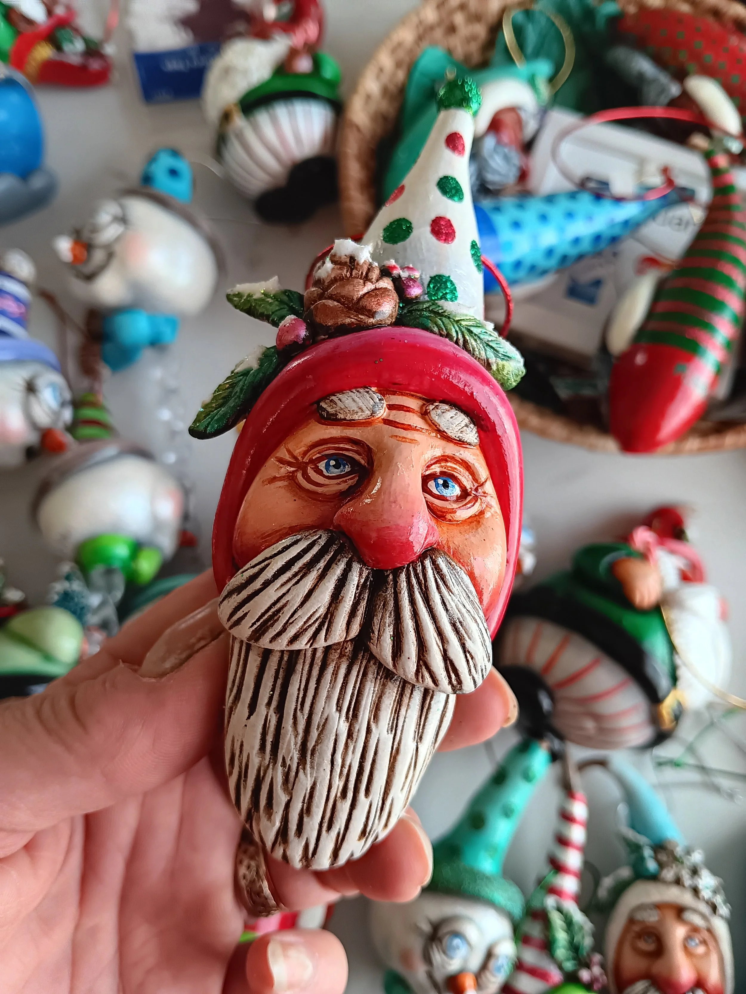 Close-up of a Christmas-themed figurine of Santa Claus with a white beard, red hat with green polka dots, and a decorative pine cone on top, held in a person's hand, with other Christmas ornaments in the background.