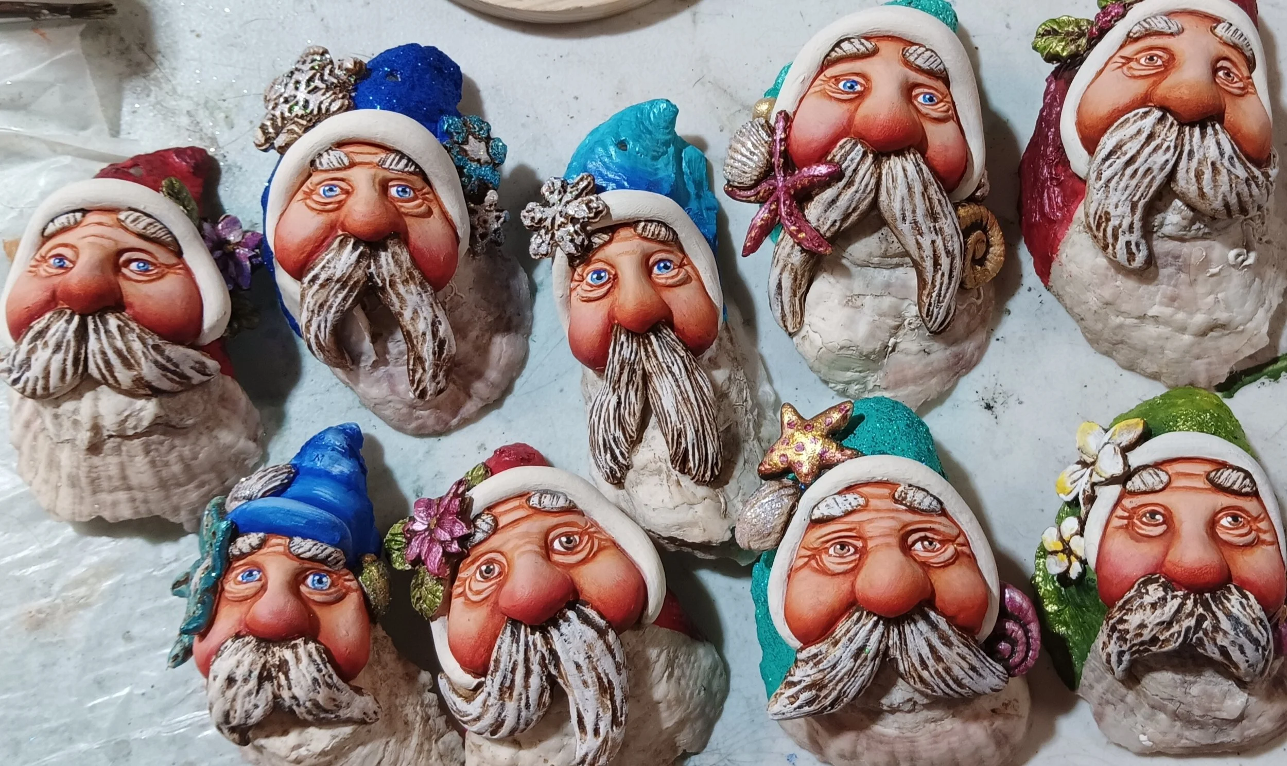 Multiple handcrafted Santa Claus figurines with colorful hats and beards, decorated with glitter and small embellishments, laid out on a surface.