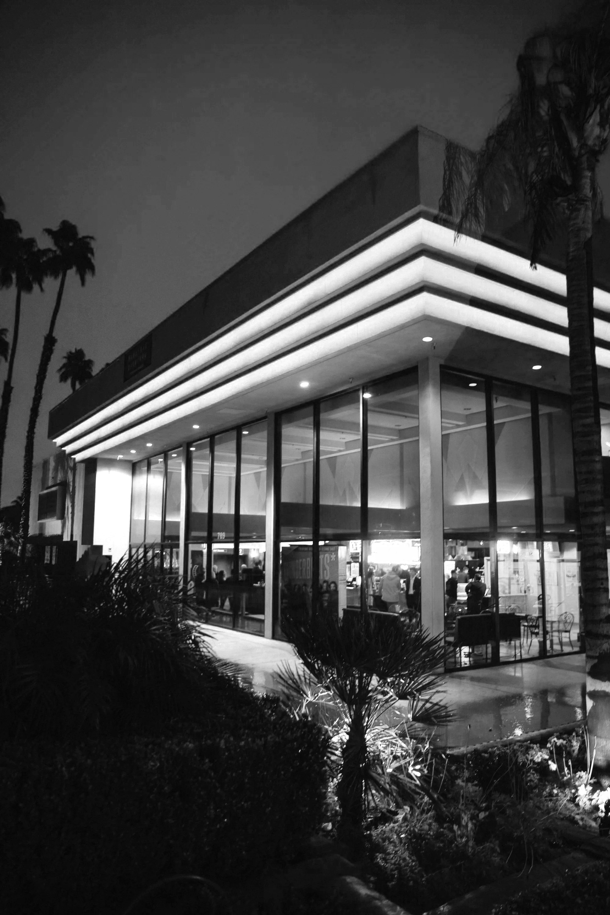 A modern, glass-fronted building illuminated at night, with palm trees nearby, possibly a restaurant or cafe.