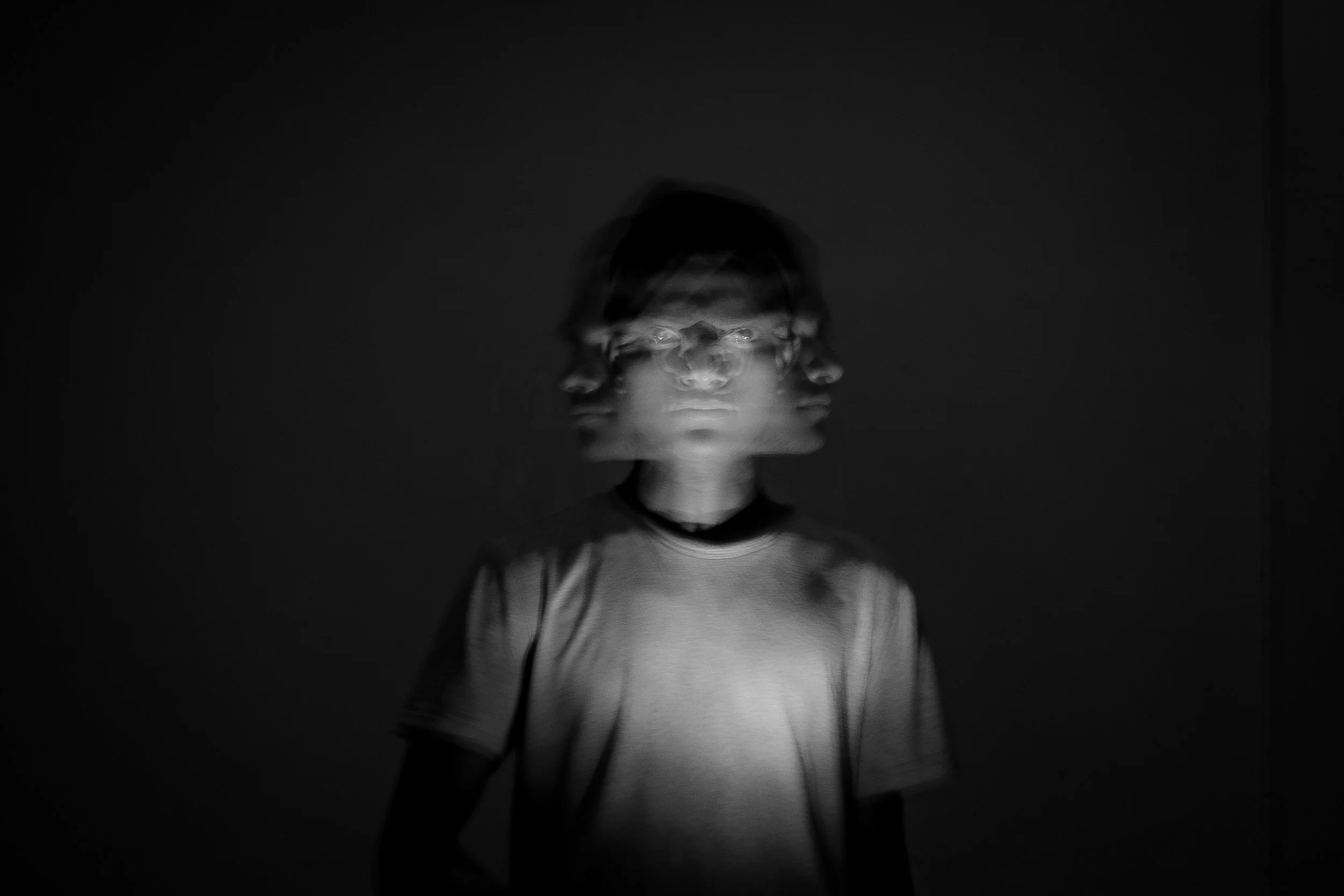 Black and white photo of a person with a blurred, multiple-exposure effect on their face, standing against a dark background.