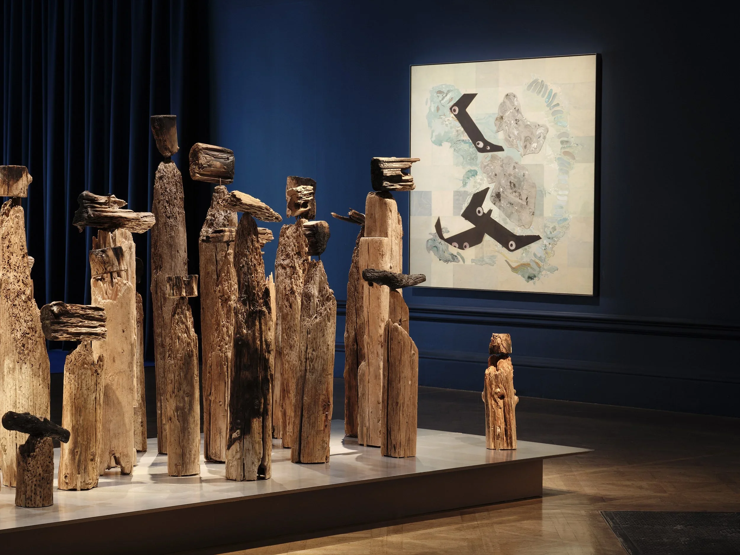 Art installation of vertically arranged weathered wood pieces of varying heights on a white platform, with dark blue curtain and an abstract painting in the background.