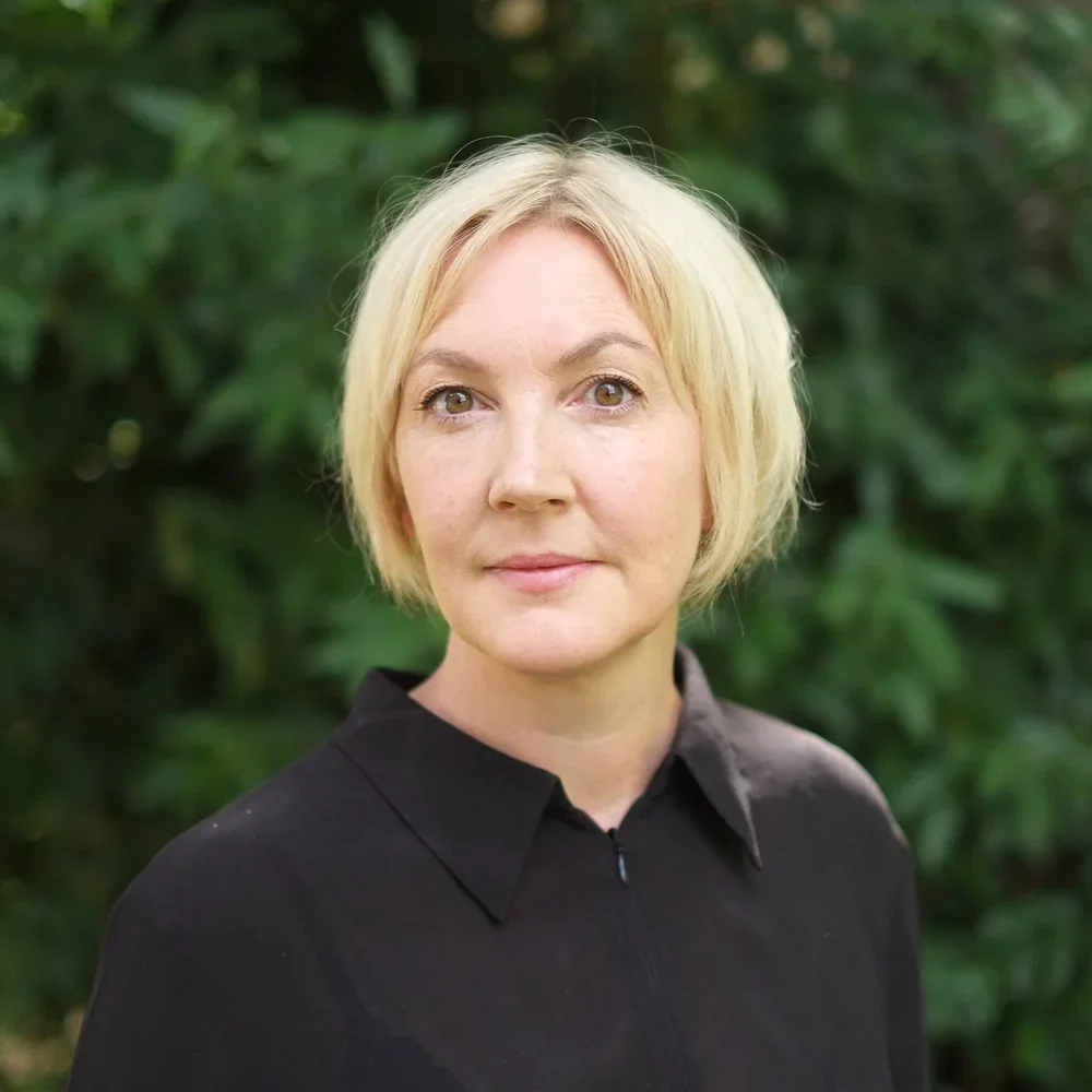 A woman with short blonde hair and light skin wearing a black shirt standing outdoors with green foliage in the background.