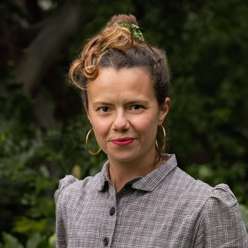 A woman with light skin and brown hair, styled in an updo with a green scrunchie, wearing gold hoop earrings and a checkered shirt, standing outdoors with green foliage in the background.