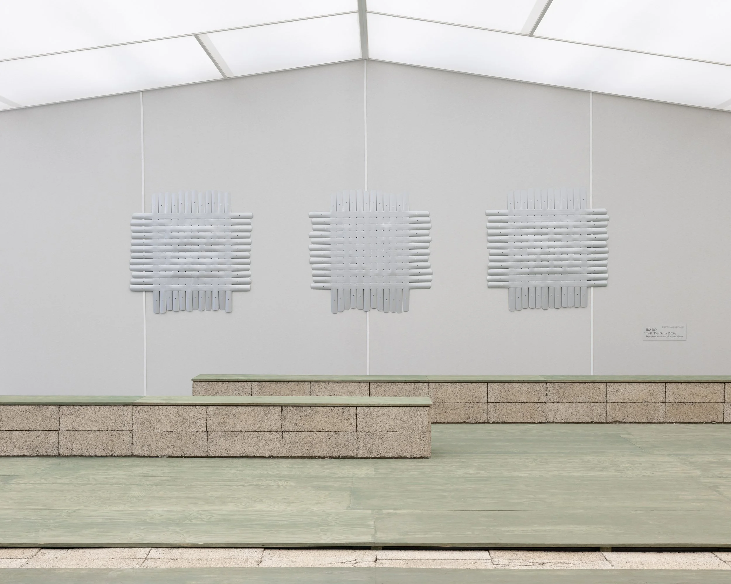 Minimalist art installation with three white, rectangular, layered sculptural pieces on a white gallery wall, accompanied by a low stone and wood bench in the foreground.