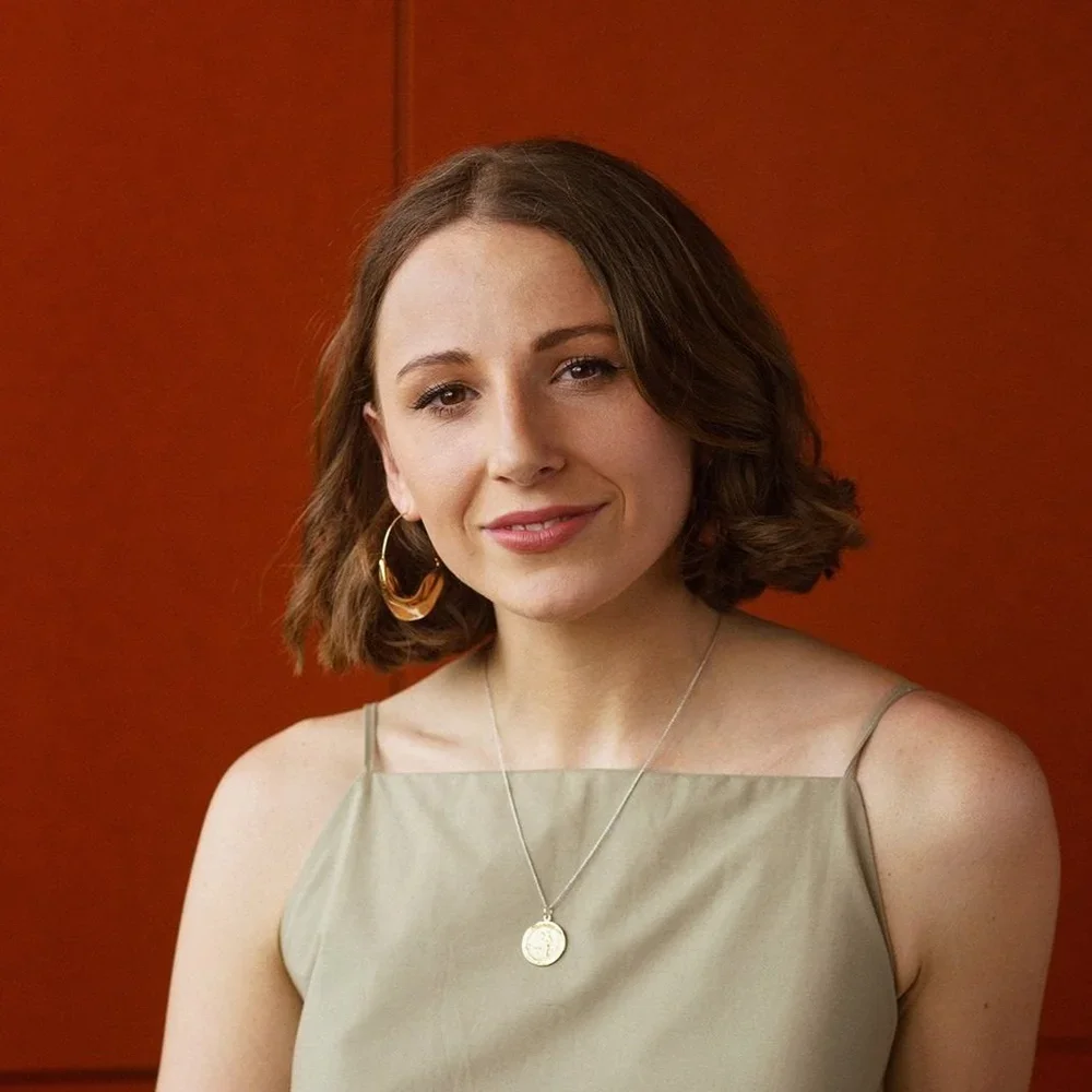 Portrait of a young woman with shoulder-length brown hair, wearing a beige sleeveless top, gold hoop earring, and a silver necklace with a round pendant, standing in front of a wooden background.