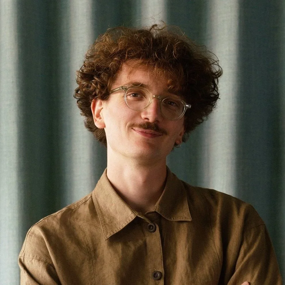 Portrait of a man with curly hair, glasses, a mustache, wearing a brown shirt, standing in front of a curtain.