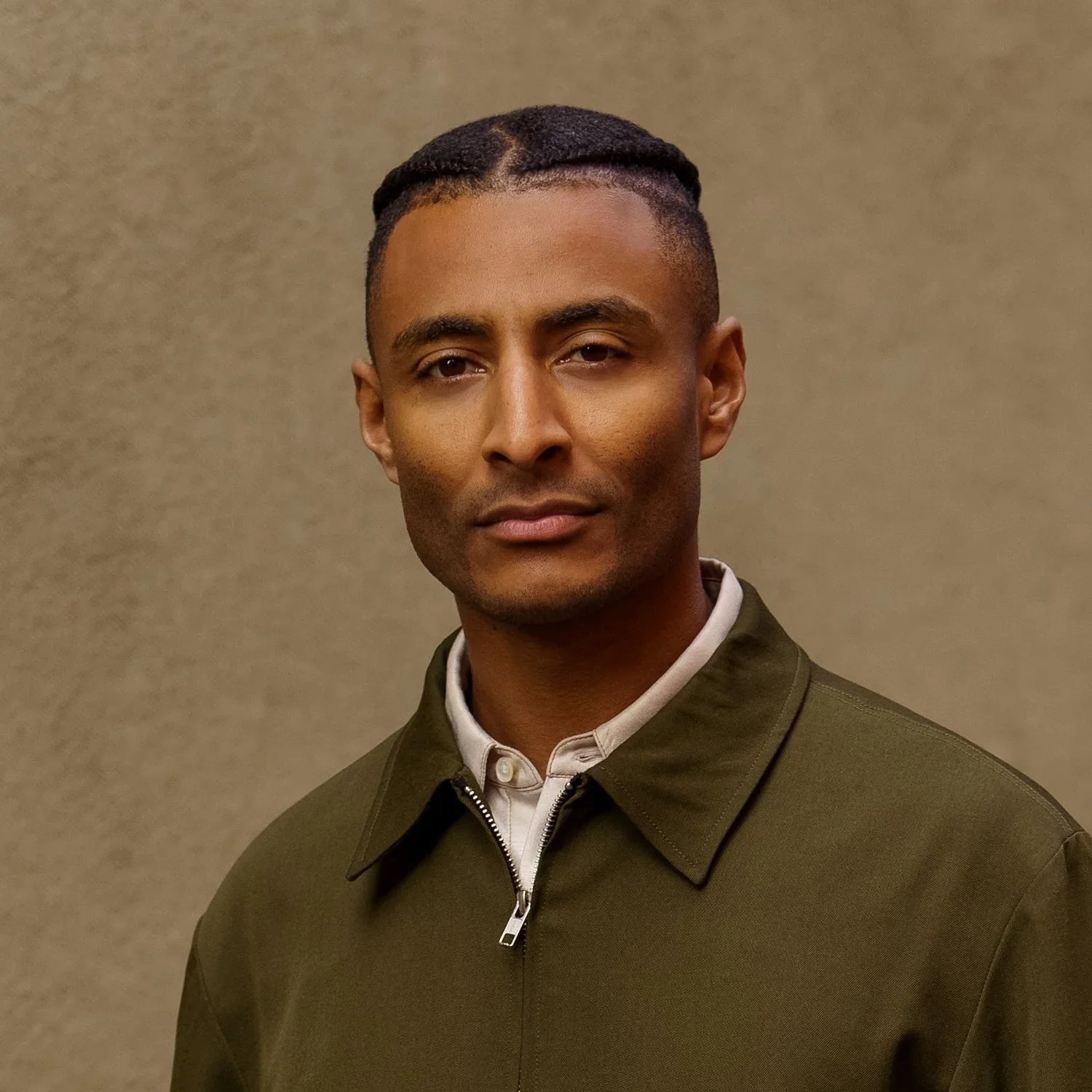 A middle-aged man with short hair styled in cornrows, wearing a dark green coat with a zipper, standing against a beige background.