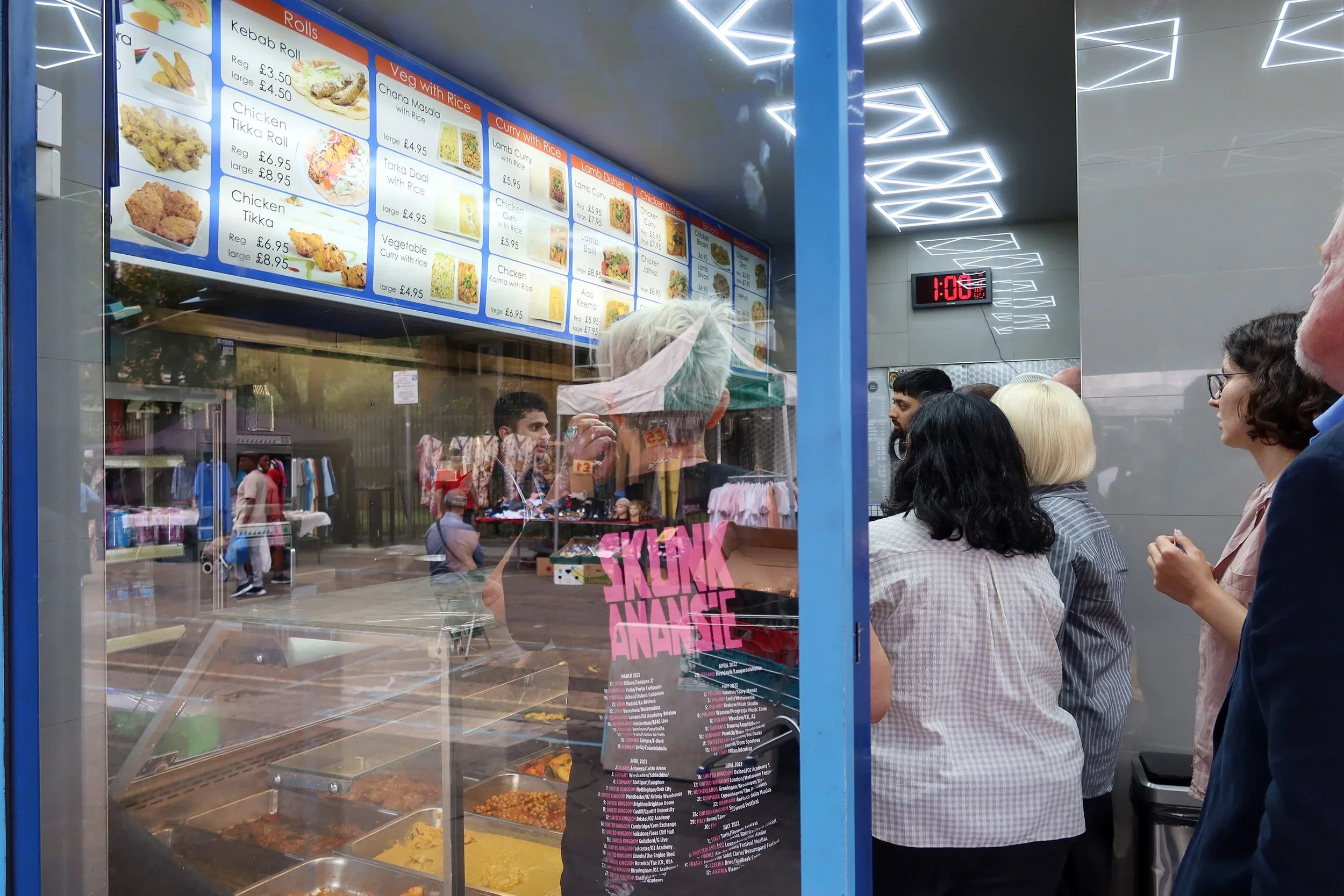 View of a busy food stall serving Indian dishes with a menu board displaying options like kebab roll, chicken tikka roll, vegetable curry, and lamb curry. Customers are waiting in line inside the stall, seen through a glass window that reflects outdoor scene with people and trees.