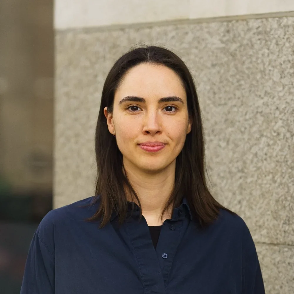 A woman with shoulder-length dark hair, wearing a navy blue shirt, standing against a beige stone wall.