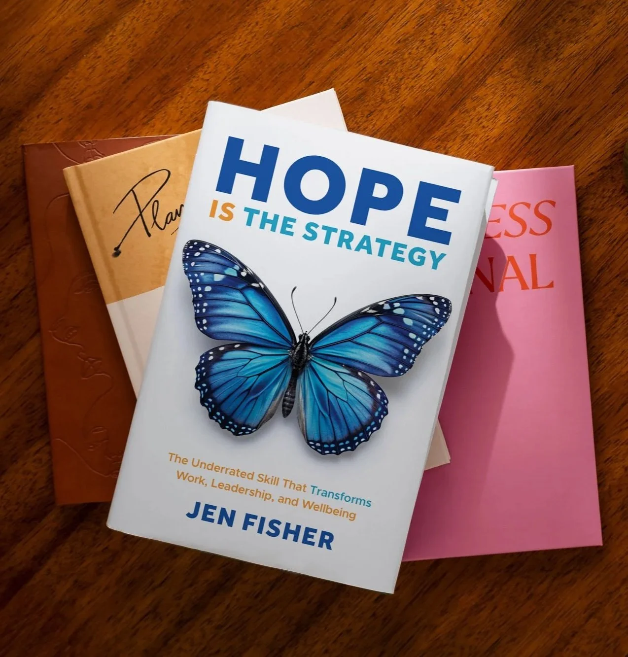 Hope is the Strategy a book with a blue butterfly on the cover sitting on a warm wooden table with other wellbeing books and journals