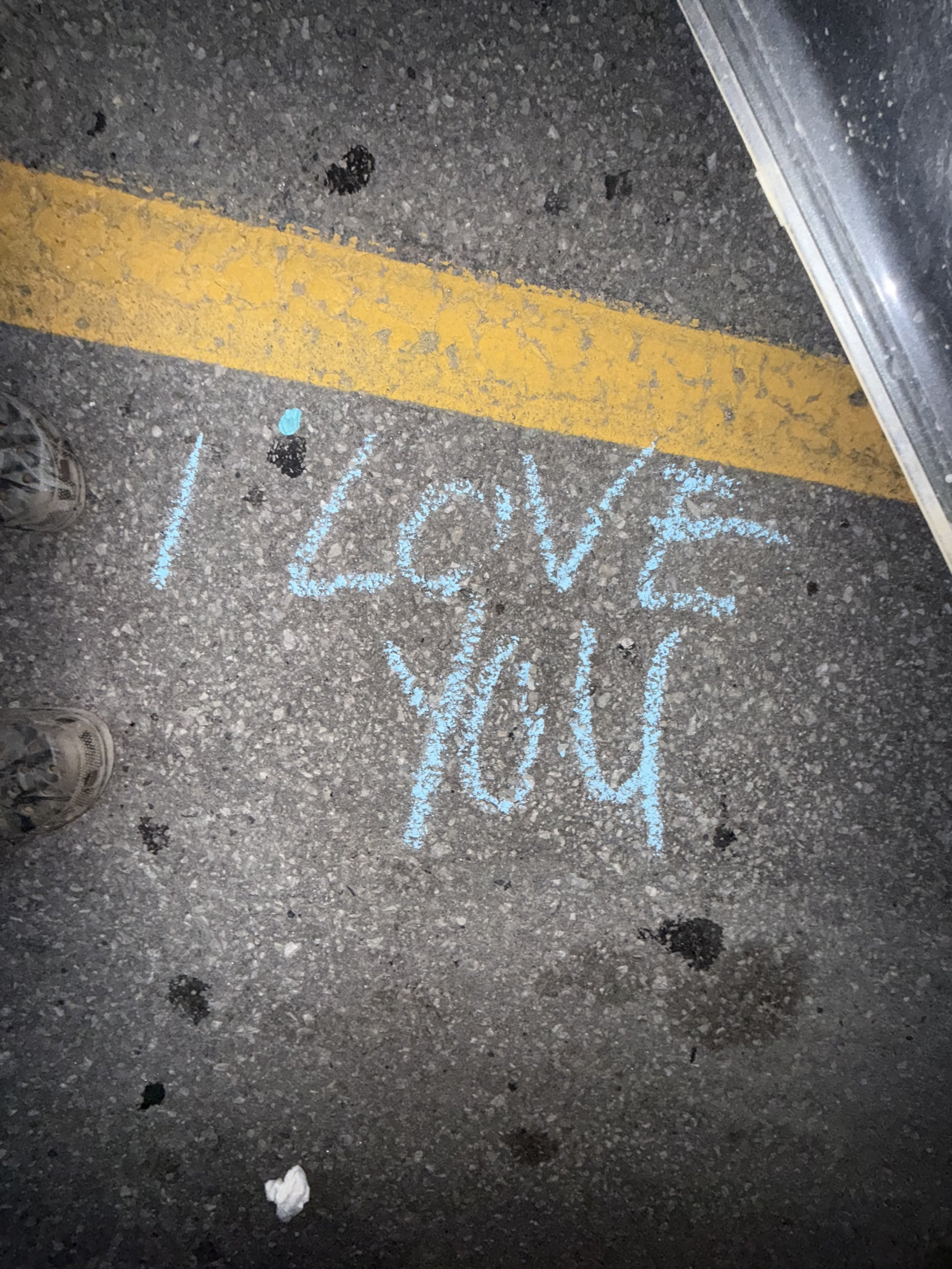 Chalk writing on asphalt that says 'I Love You' with a yellow parking lot line and shadows of shoes and part of a vehicle.